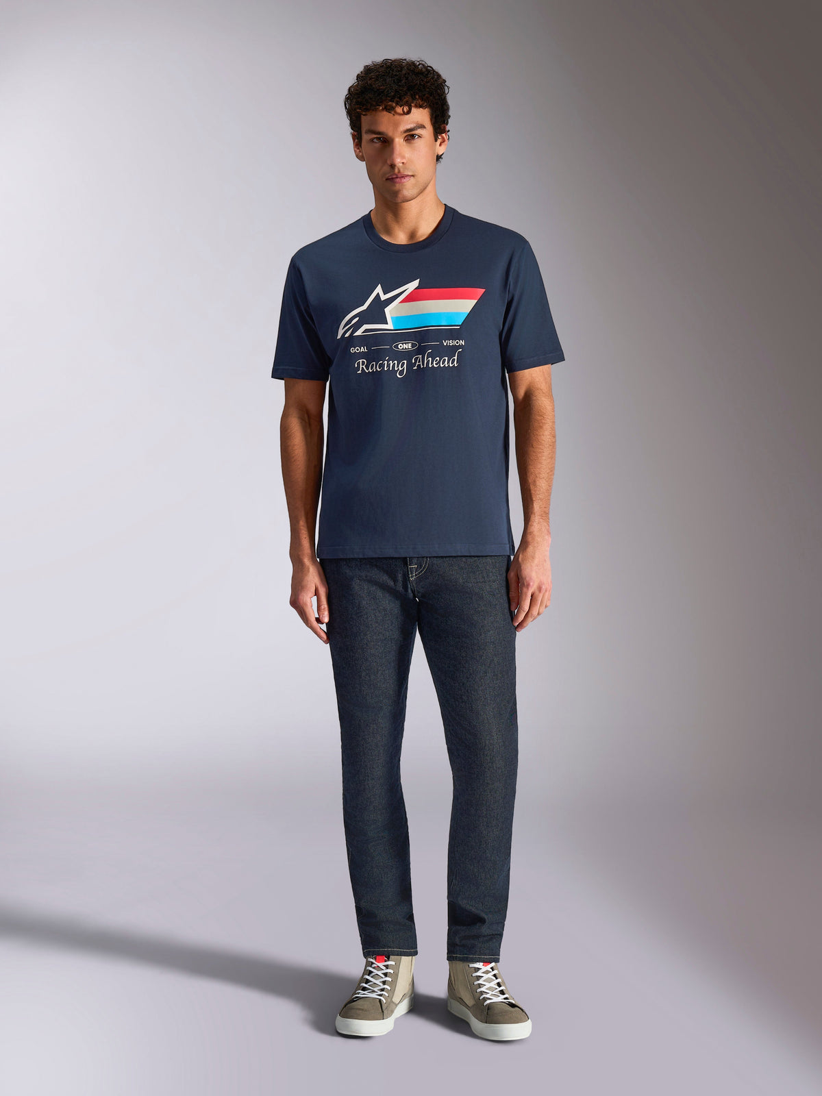 Camiseta Alpinestars Trifecta CSF, camiseta de manga corta, azul marino, logo Astars blanco con rayas rojas, blancas y azul claro y texto 'Racing Ahead', combinada con vaqueros oscuros y zapatillas verde oliva, estilo streetwear casual de automovilismo