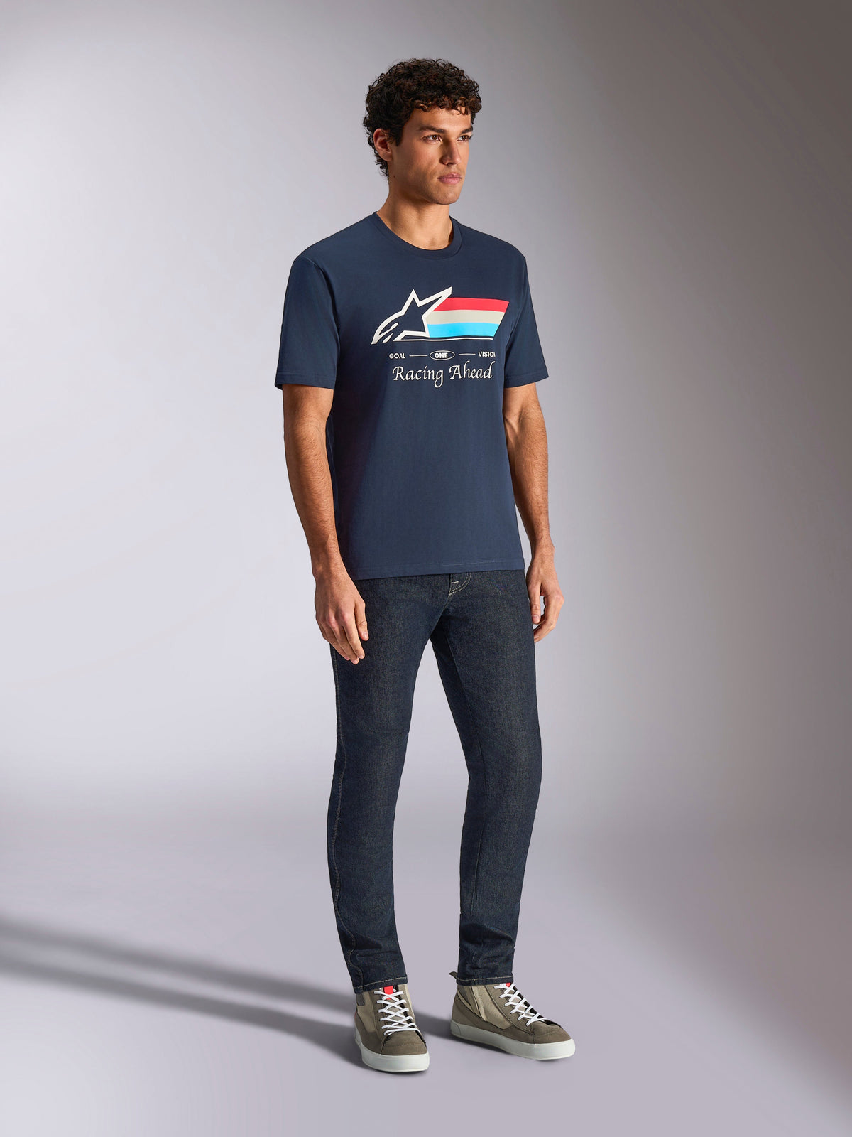 Alpinestars Trifecta CSF Tee, camiseta de manga corta, azul marino, con el logotipo de Astars con franjas tricolores y el texto Racing Ahead, usada por un modelo con vaqueros oscuros y zapatillas, ropa urbana casual inspirada en el automovilismo