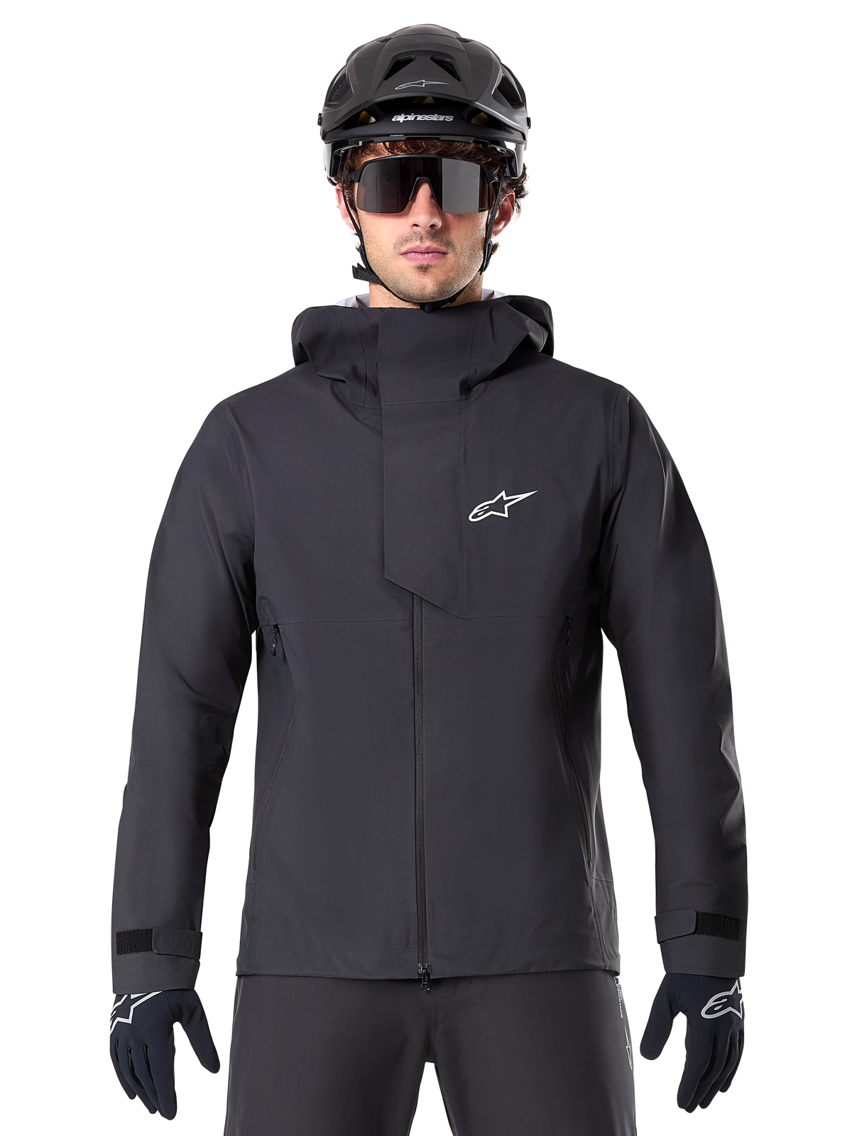 Chaqueta impermeable Alpinestars A-Dura Elite, negra, chaqueta de ciclismo de montaña, diseño de cuello alto con capucha, cremallera frontal con solapa protectora en el pecho y logotipo blanco, usada con casco negro y guantes de ciclismo, protección técnica contra la intemperie para rodar por senderos