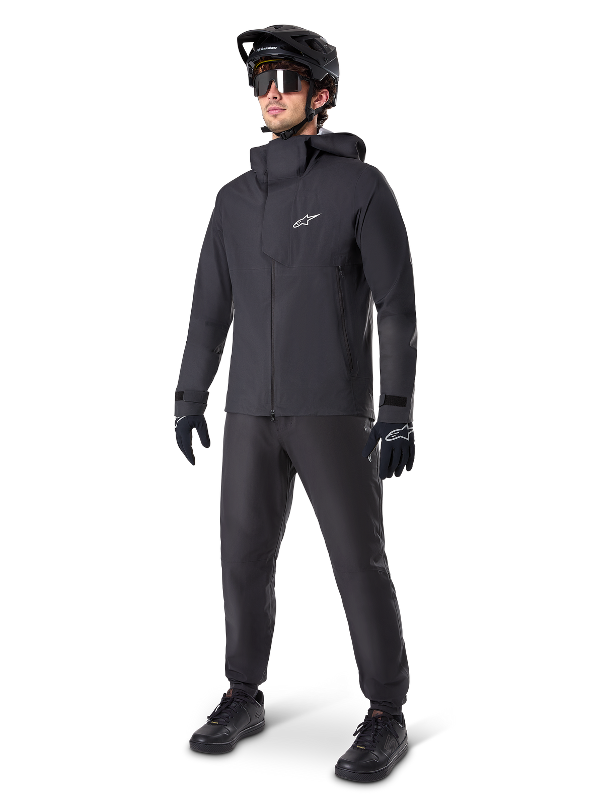 Chaqueta impermeable Alpinestars A-Dura Elite en color negro, usada por un ciclista con pantalones de MTB, guantes y casco negros a juego. Presenta un diseño técnico impermeable con el logotipo Astars blanco en el pecho y un cuello protector alto.