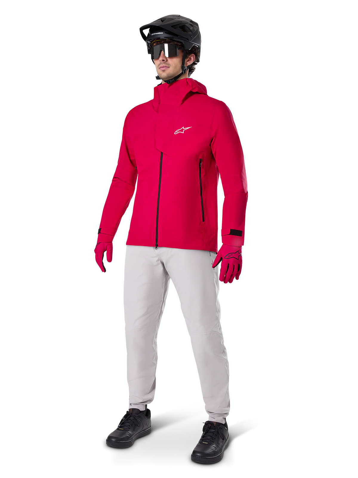 Ciclista masculino con la chaqueta impermeable Alpinestars A-Dura Elite en rojo flúor, combinada con pantalones técnicos gris claro, guantes rojos, casco de MTB negro con gafas y zapatillas negras, equipamiento completo de alto rendimiento para ciclismo de montaña