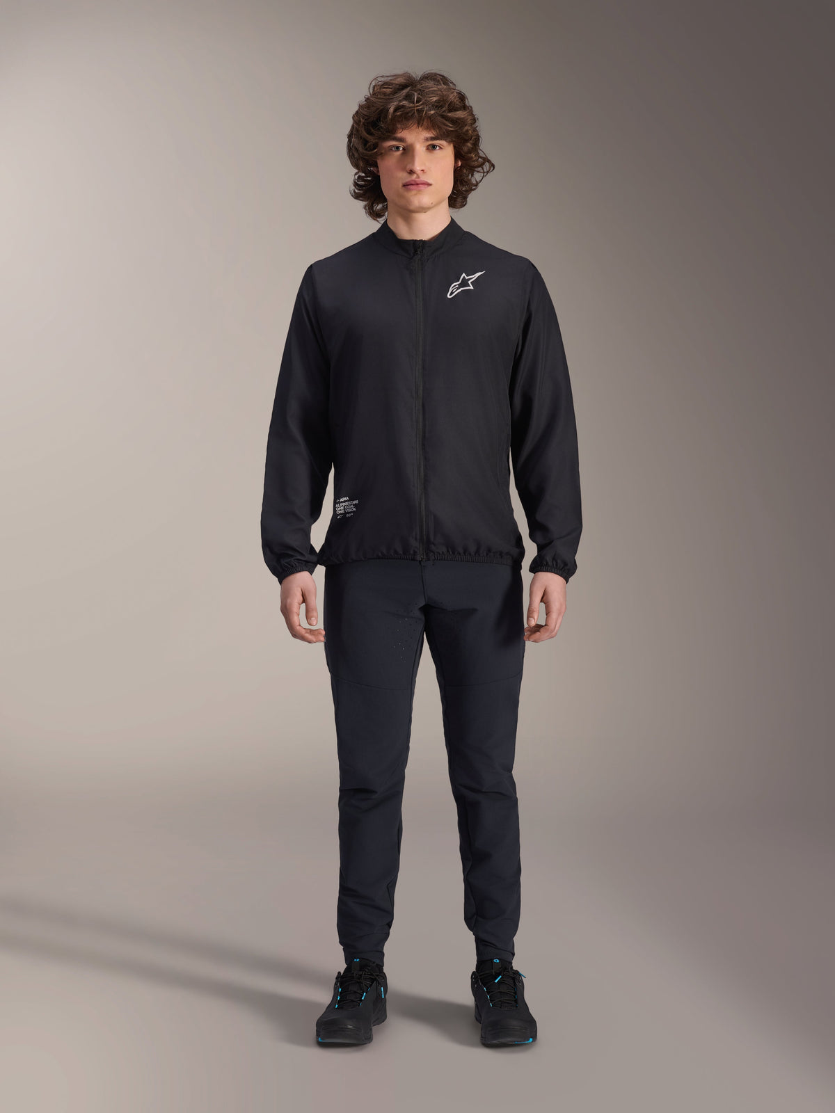 Modelo con la chaqueta cortavientos Alpinestars A-Aria en color negro, con cremallera frontal completa, cuello simulado y logotipo Astars blanco en el pecho, combinada con pantalones técnicos negros y calzado negro, ropa de ciclismo de rendimiento para ciclismo de montaña y uso al aire libre.