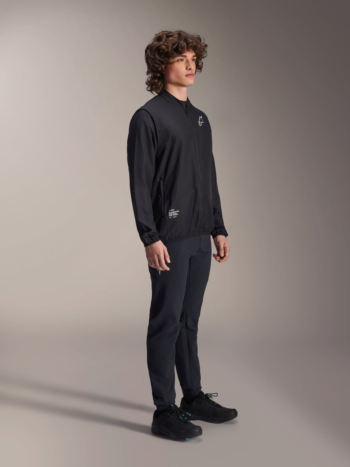 Chaqueta Cortavientos Alpinestars A-Aria, negra, chaqueta de ciclismo ligera y resistente al viento lucida por un modelo con pelo rizado, con un logotipo blanco de Astars en el pecho y marca en la parte inferior derecha, combinada con pantalones y calzado negros, ajuste de rendimiento para mountain bike y gravel