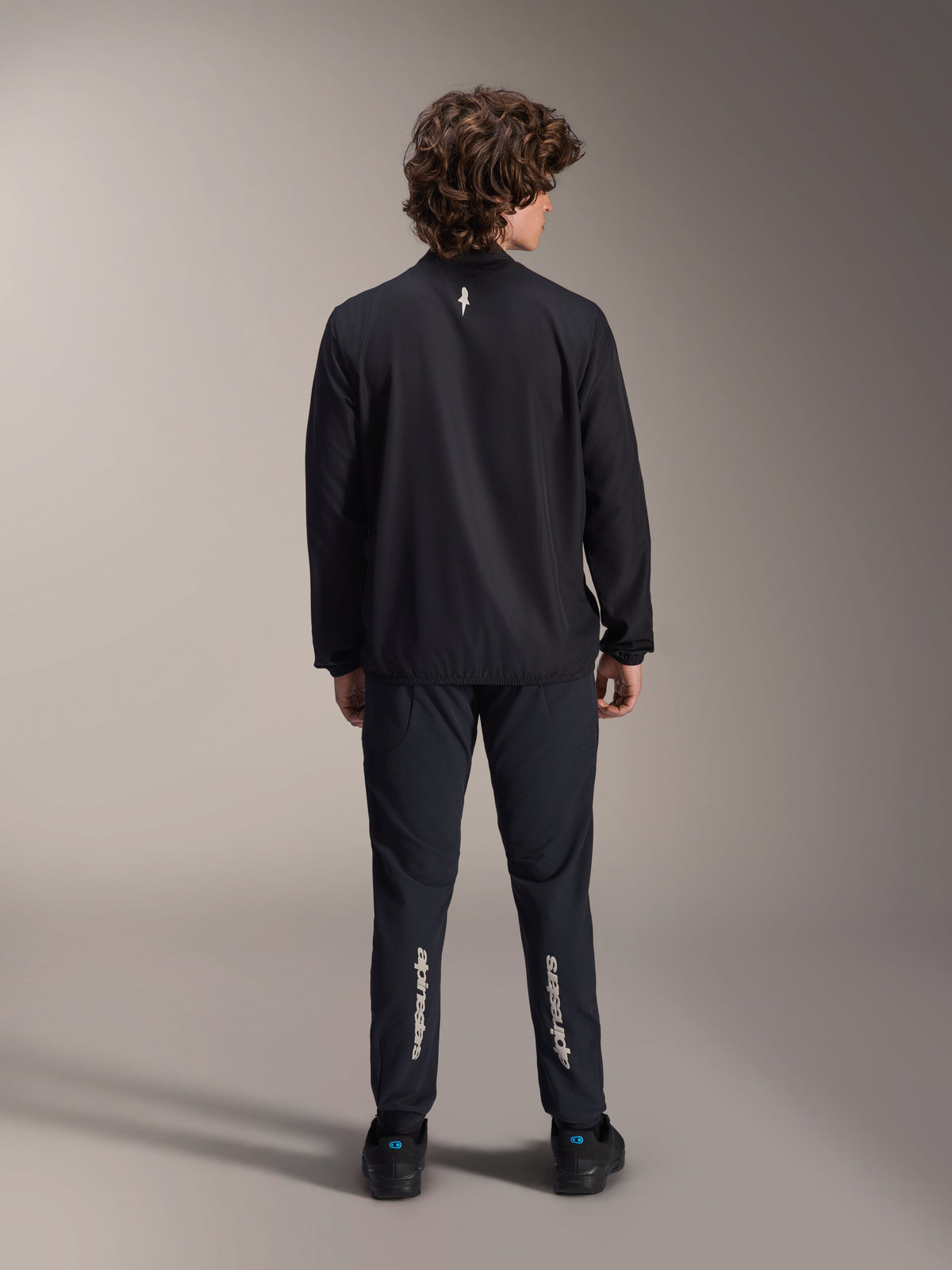Chaqueta cortavientos Alpinestars A-Aria, negra, vista trasera, chaqueta de ciclismo ligera y resistente al viento con un pequeño logotipo blanco en la parte superior de la espalda, combinada con pantalones negros a juego con la marca Alpinestars vertical en las pantorrillas, equipamiento de alto rendimiento para bicicleta de montaña