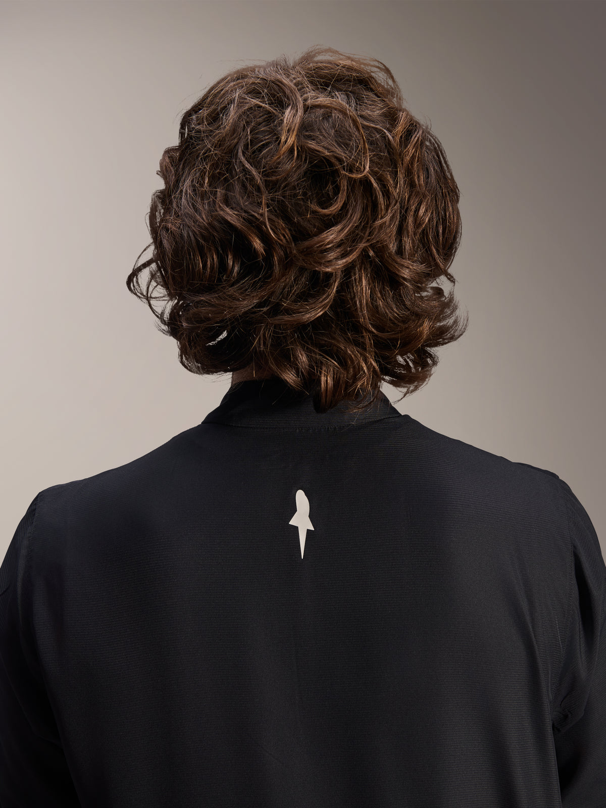 Chaqueta cortavientos Alpinestars A-Aria, negra, primer plano de la parte posterior de los hombros y el cuello, con un pequeño logotipo blanco en el centro de la espalda, usada por un modelo con cabello castaño ondulado, equipamiento de ciclismo de rendimiento minimalista
