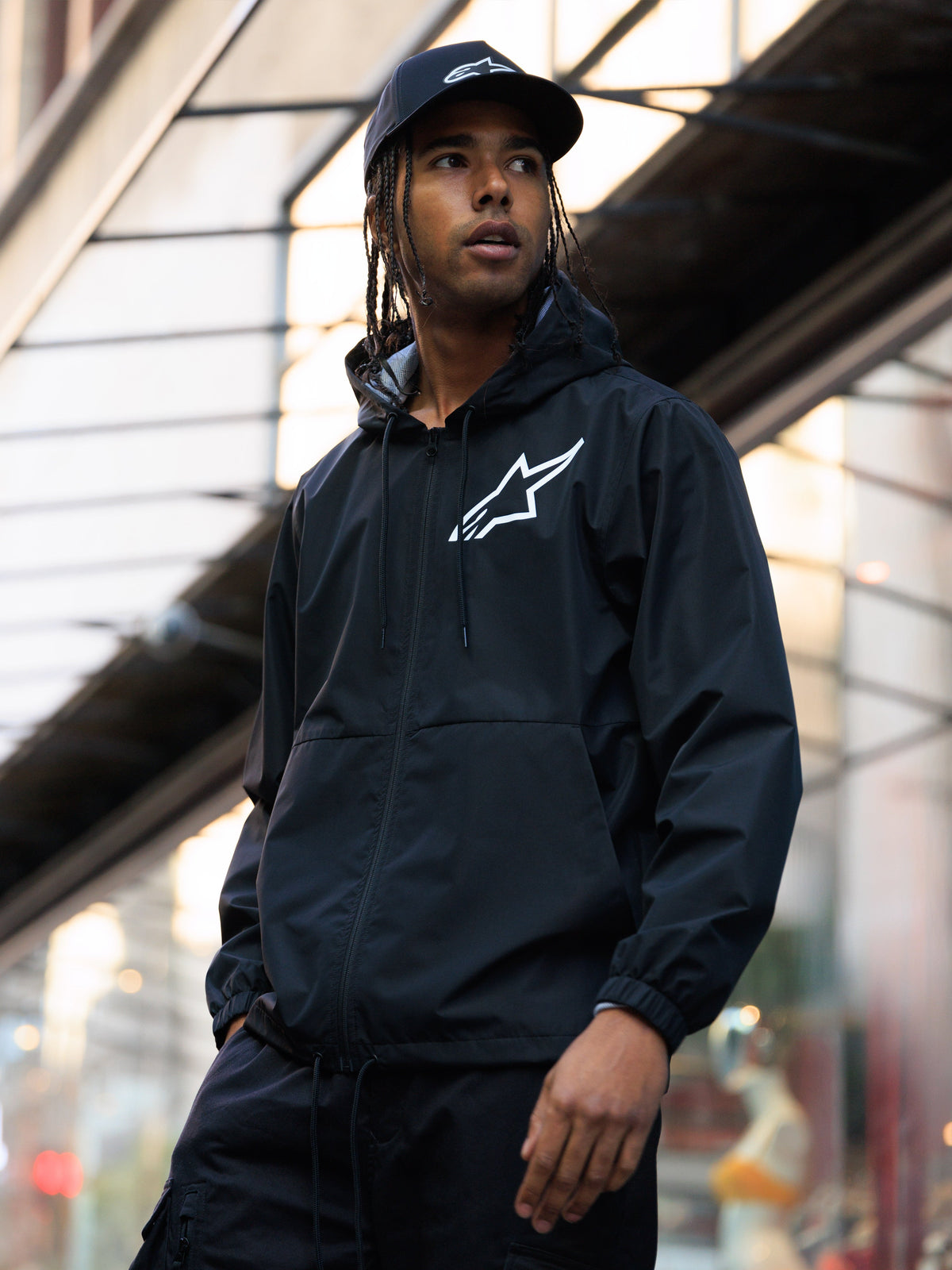 Chaqueta cortavientos Alpinestars Speeder Chromium, negra, cortavientos con capucha con un logotipo blanco en el pecho, usada por un hombre con una gorra Alpinestars negra a juego en un entorno urbano, estilo casual de streetwear motorsport.