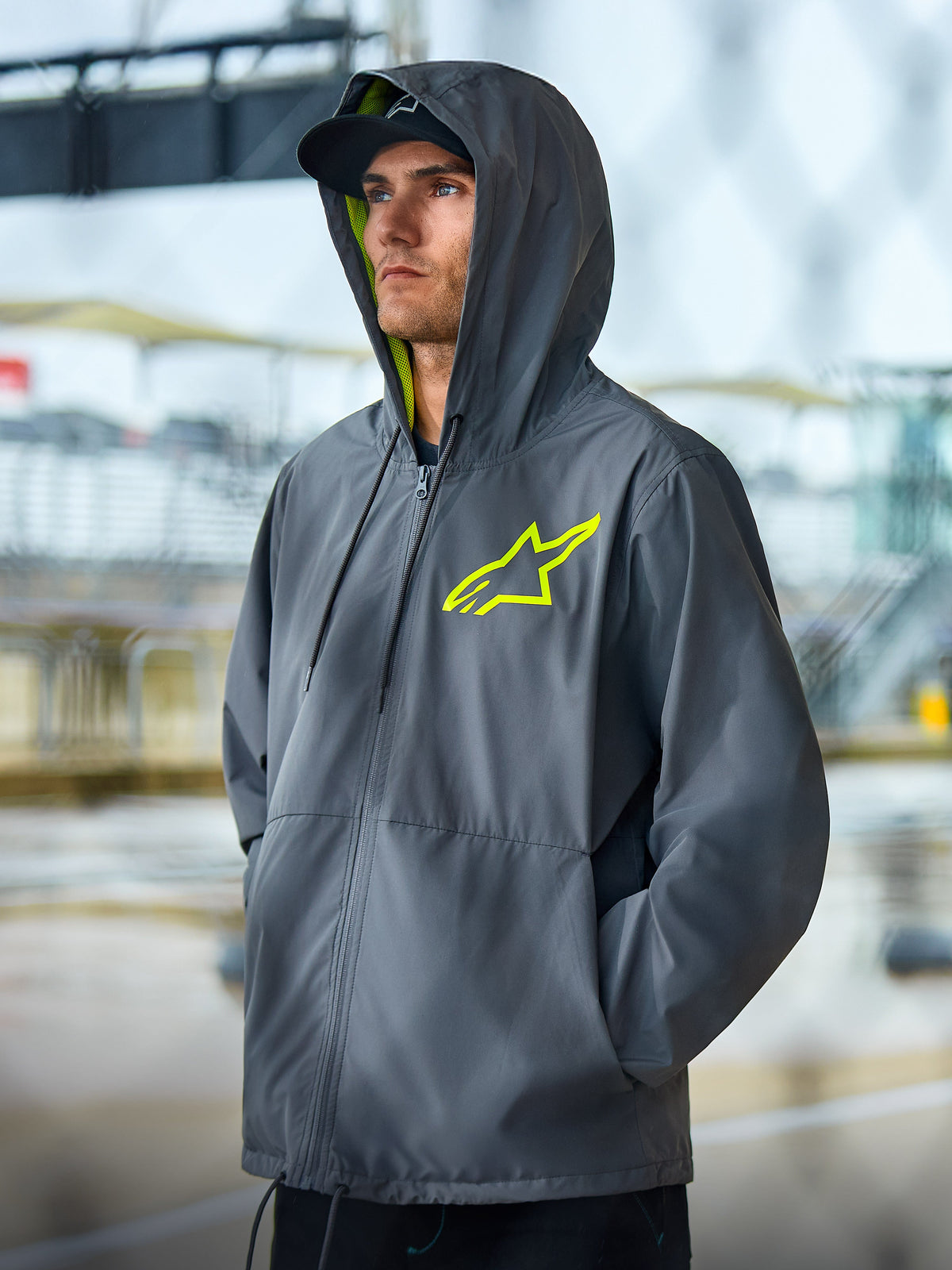 Alpinestars Speeder Chromium Windbreak Jacket, Charcoal grey lifestyle jacket with high-visibility yellow accents, hooded design featuring yellow interior lining and chest logo, casual motorsport streetwear worn by a model in an outdoor setting