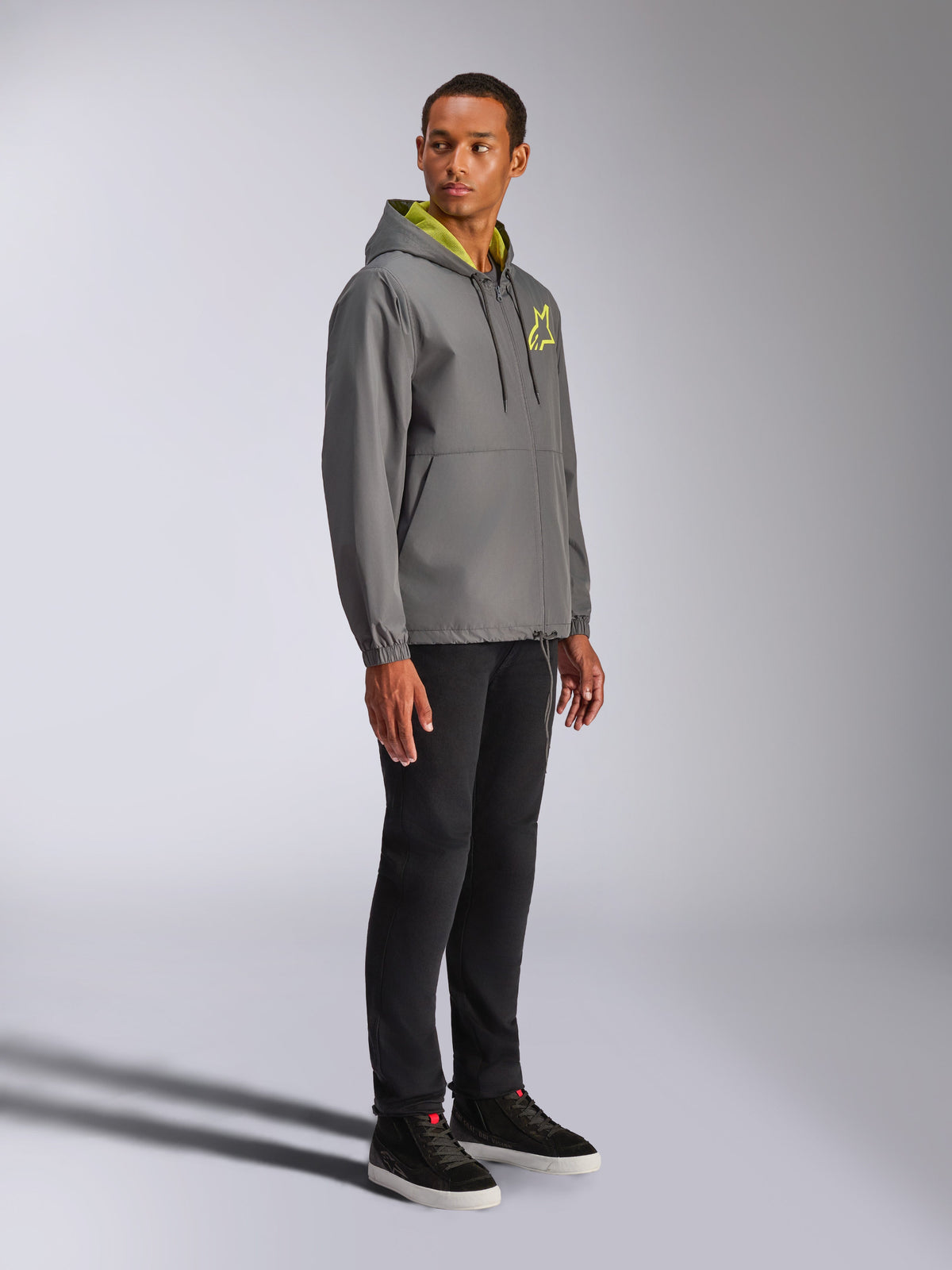 Alpinestars Speeder Chromium Windbreak Jacket, Charcoal with Hi-Vis Yellow accents, male model wearing hooded windbreaker, yellow star logo on chest, paired with black pants and black sneakers, casual motorsport-inspired streetwear
