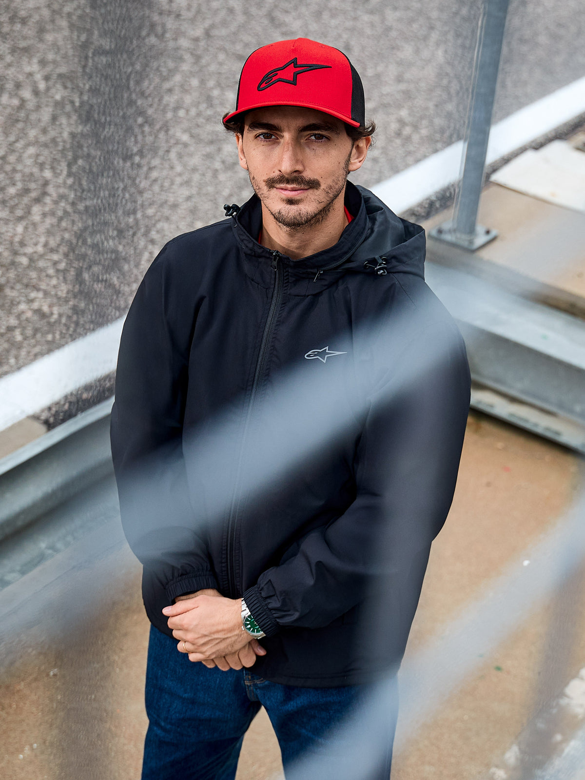 Hombre luciendo la chaqueta Overhaul Racer de Alpinestars en color negro, una chaqueta de lluvia con capucha y tejido resistente al agua con un pequeño logotipo en el pecho, complementada con una gorra roja de Alpinestars, ropa casual de estilo de vida motorsport