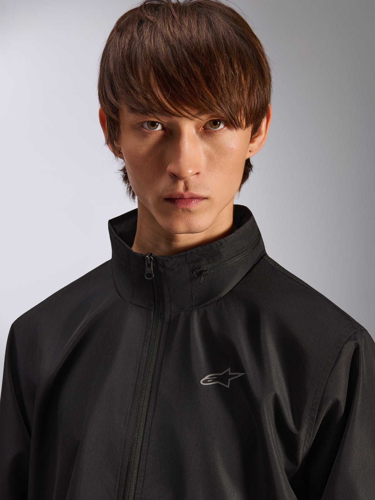 Chaqueta Alpinestars Overhaul Racer, chaqueta impermeable negra, primer plano de un modelo masculino con la chaqueta, con cuello alto y pequeño logo plateado de Astars en el pecho, prenda exterior informal inspirada en el automovilismo