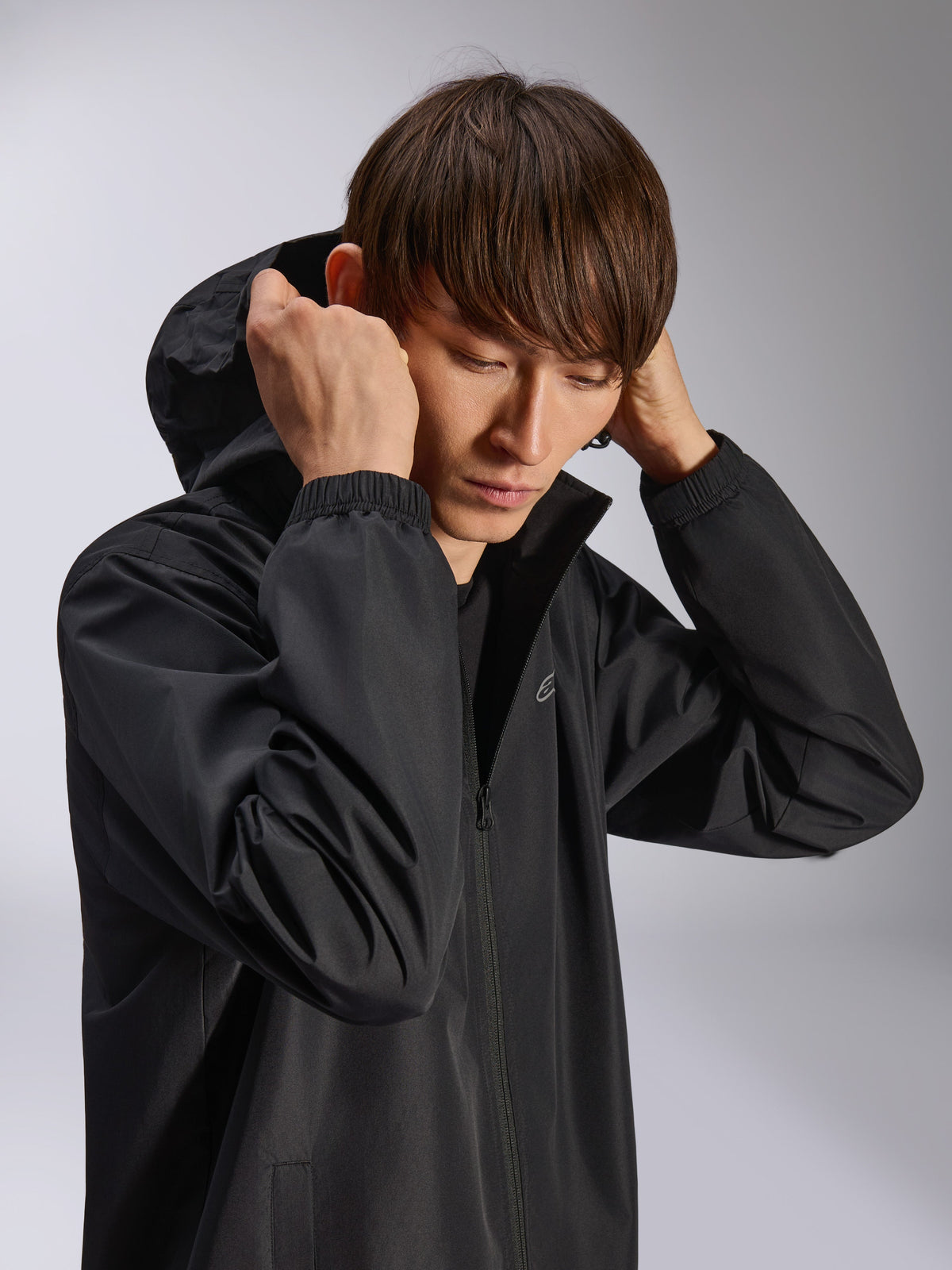 Chaqueta Alpinestars Overhaul Racer, chaqueta impermeable negra con capucha, usada por un modelo con la capucha puesta, con tejido resistente al agua de 2.5L, puños elásticos y logotipo minimalista en el pecho, prenda técnica casual inspirada en el automovilismo