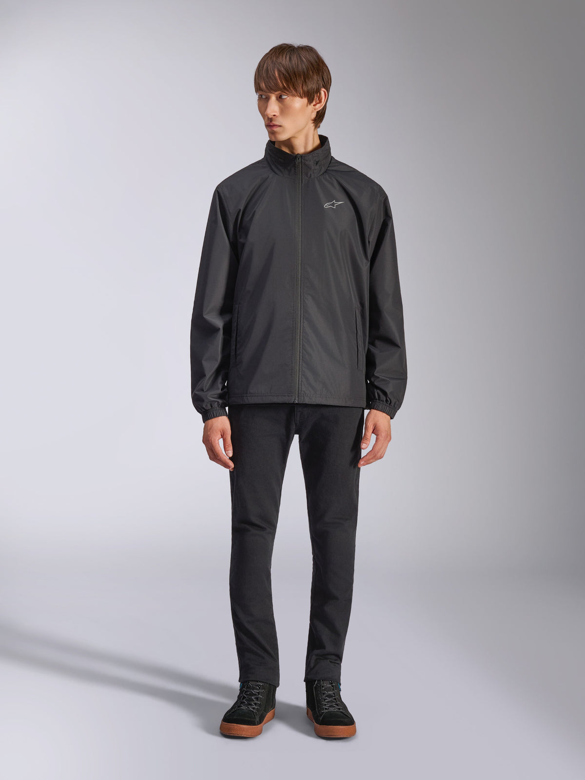 Chaqueta Overhaul Racer de Alpinestars, chaqueta de lluvia negra para hombre, vista de cuerpo completo en un modelo con pantalones negros y zapatillas casuales, con un pequeño logotipo blanco en el pecho, tejido resistente al agua y diseño informal inspirado en el automovilismo