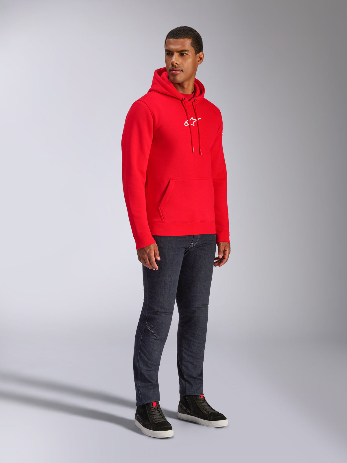 Sudadera con capucha Alpinestars Frontal, roja, modelo masculino que viste sudadera de forro polar con logotipo de estrella A blanco en el pecho, bolsillo tipo canguro, capucha con cordón, combinada con vaqueros de denim oscuro, ropa urbana casual inspirada en el automovilismo