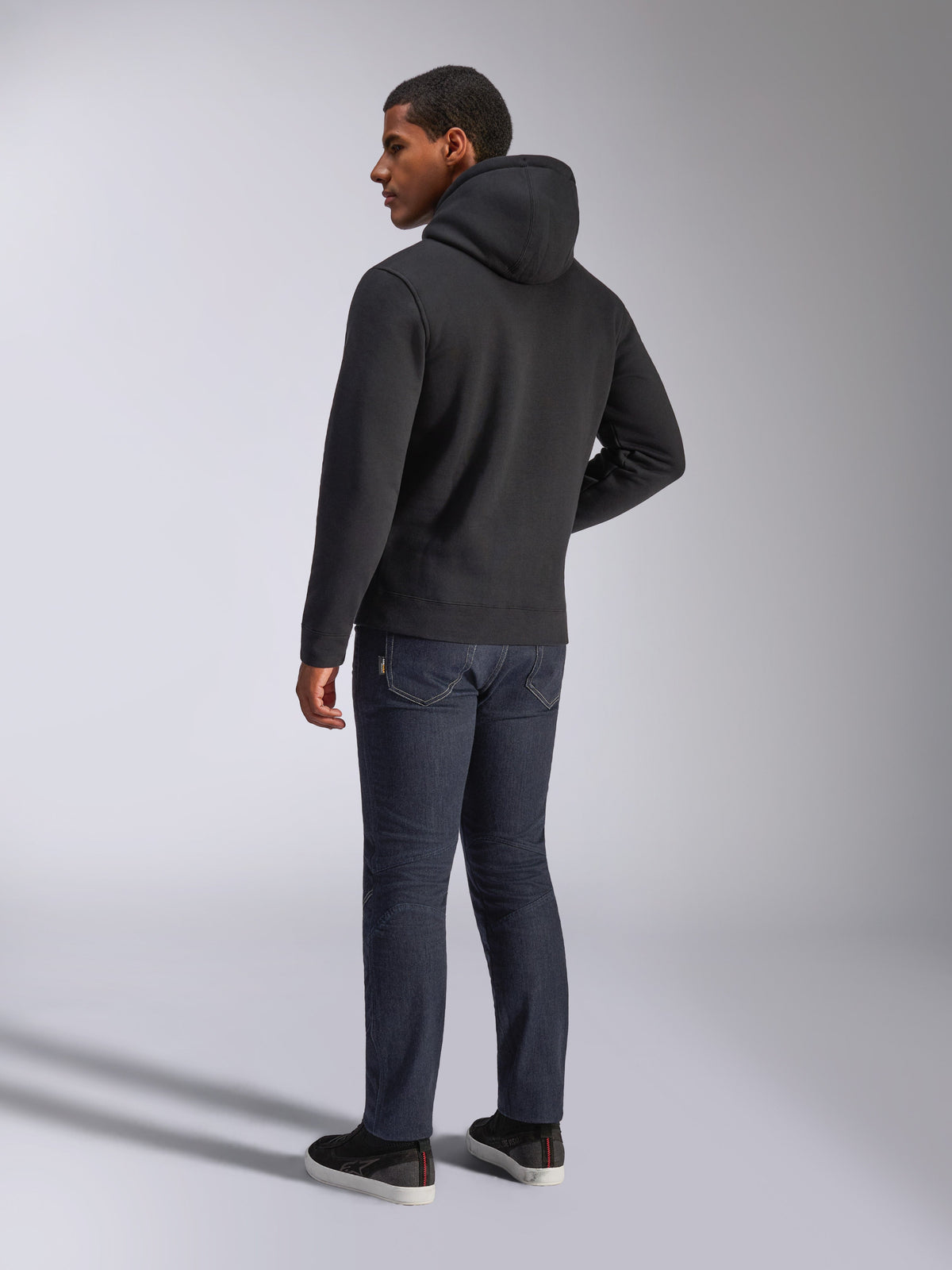 Alpinestars Blaze V3 Hoodie, back view of a man wearing the black pullover hoodie, paired with dark jeans and black sneakers, casual motorsport-inspired streetwear