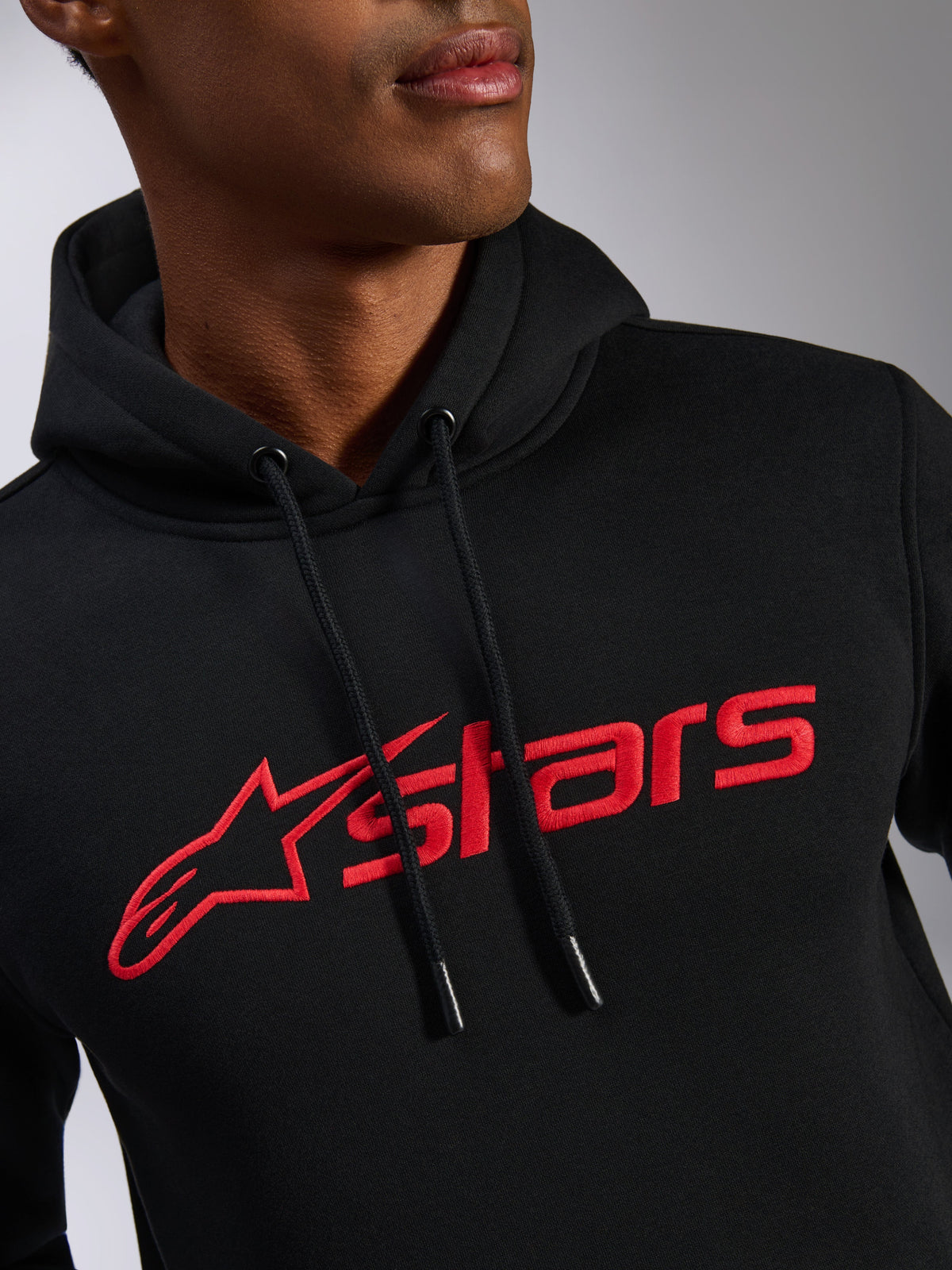 Alpinestars Blaze V3 Hoodie, close-up view of the chest, black fleece fabric with red Alpinestars linear logo print, drawstring hood with dipped tips, casual motorsport-inspired streetwear