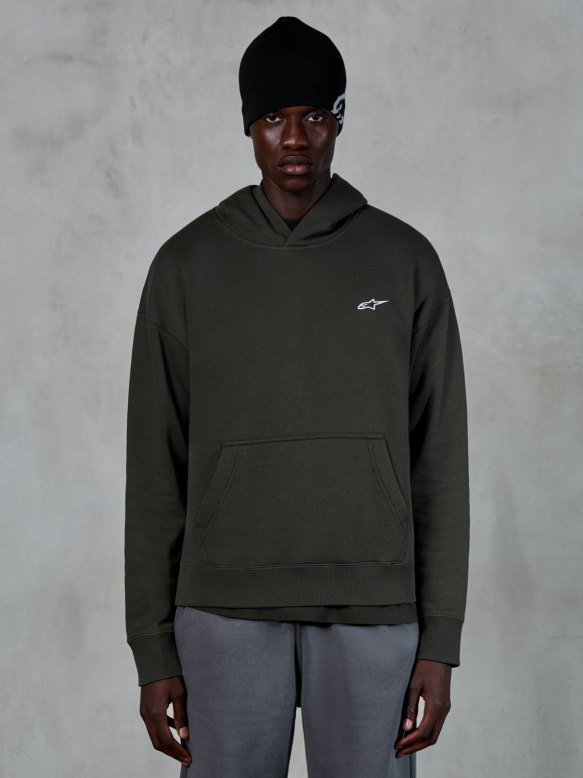Sudadera con capucha Alpinestars Essentials Ageless, estilo streetwear casual, pullover en color Off Black con un pequeño logo de Alpinestars en blanco en el pecho, usada con un gorro negro, diseño minimalista con bolsillo de canguro delantero y acabado lavado a la piedra