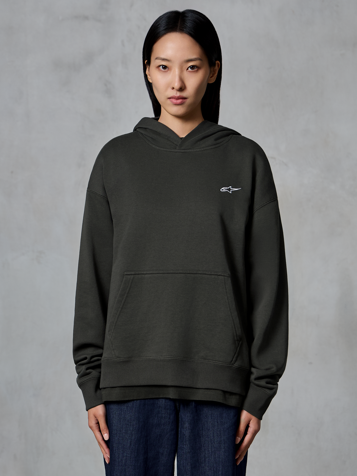 Alpinestars Essentials Ageless Hoodie, negro (Off Black), usada por una modelo, con un pequeño logo blanco de Astars en el pecho, bolsillo de canguro frontal, acabado lavado a la piedra, estilo urbano de forro polar premium