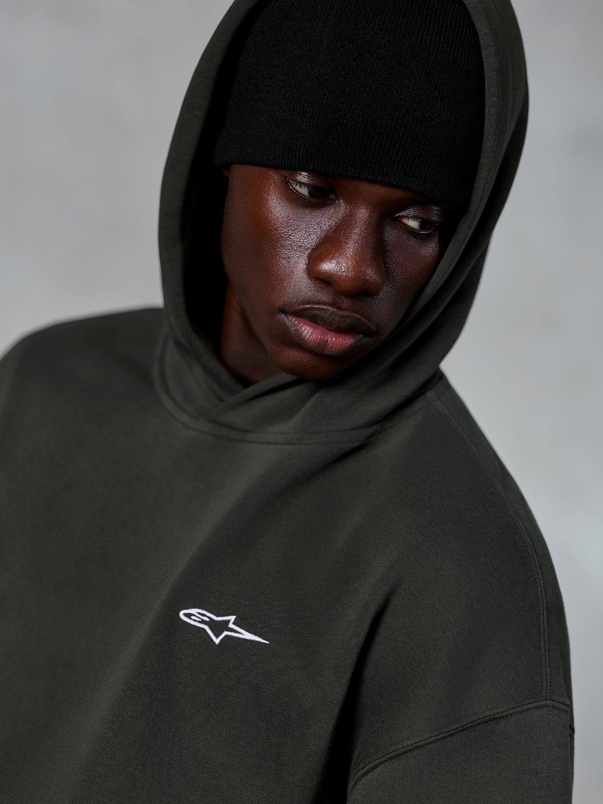 Alpinestars Essentials Ageless Hoodie, Off Black, primer plano de un modelo con un gorro negro debajo de la capucha, con un pequeño logotipo de estrella blanca en el pecho, ropa urbana casual inspirada en el motociclismo