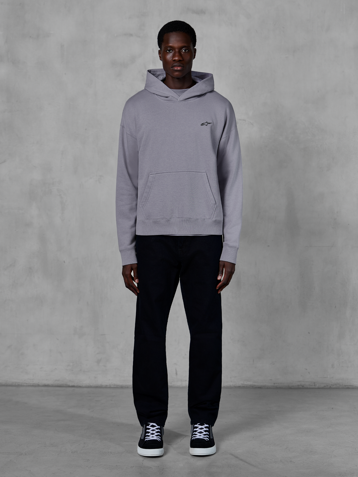 Alpinestars Essentials Ageless Hoodie, Silver, male model wearing casual streetwear, small black Alpinestars logo on chest, front kangaroo pocket, minimalist design, paired with black pants and sneakers.