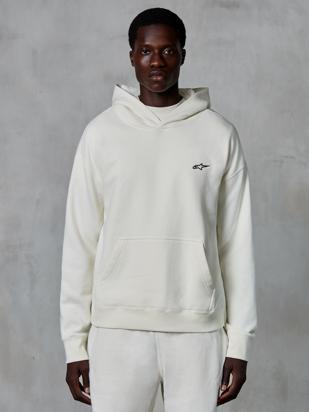 Alpinestars Essentials Ageless Hoodie, color blanco roto, usada por modelo masculino, pequeño logotipo negro en el pecho, bolsillo tipo canguro frontal, manga larga, ropa urbana casual inspirada en el automovilismo