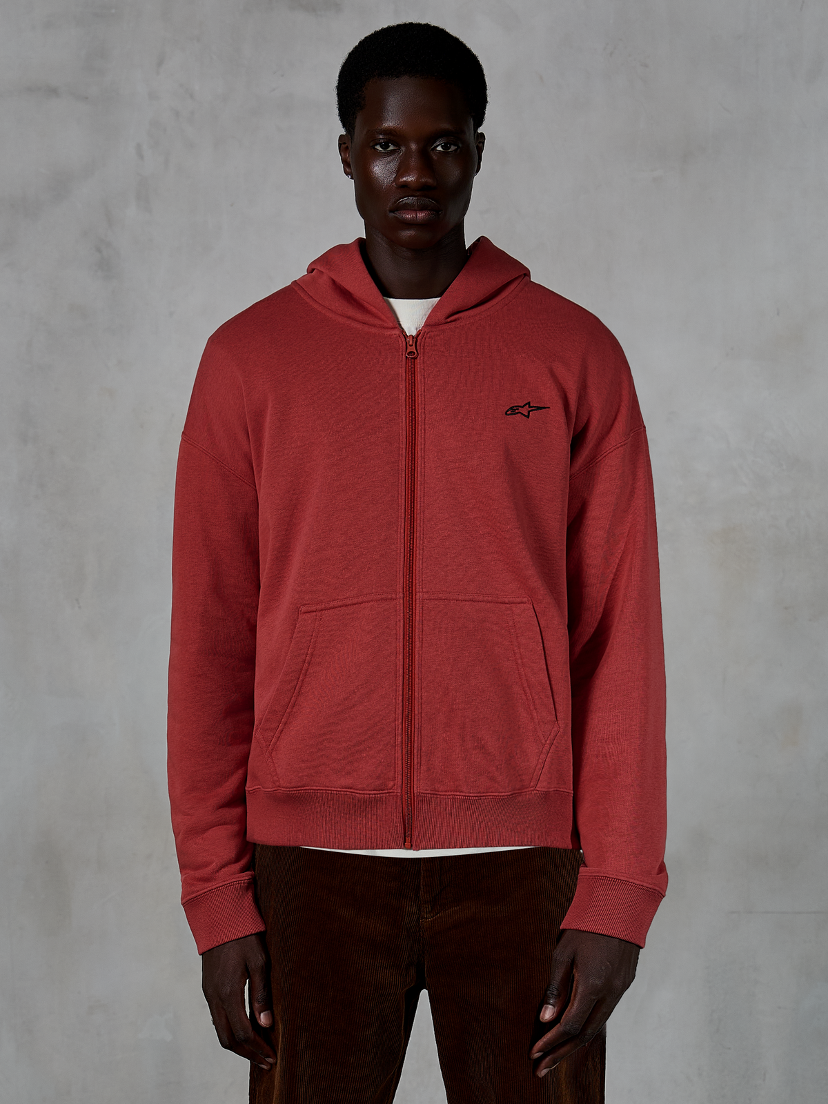 Sudadera con capucha y cremallera Alpinestars Essentials Ageless, sudadera con capucha y cremallera completa en color rojo ladrillo, lucida por un modelo con pantalones de pana marrones, con un pequeño logotipo negro en el pecho, estilo urbano informal de ajuste relajado