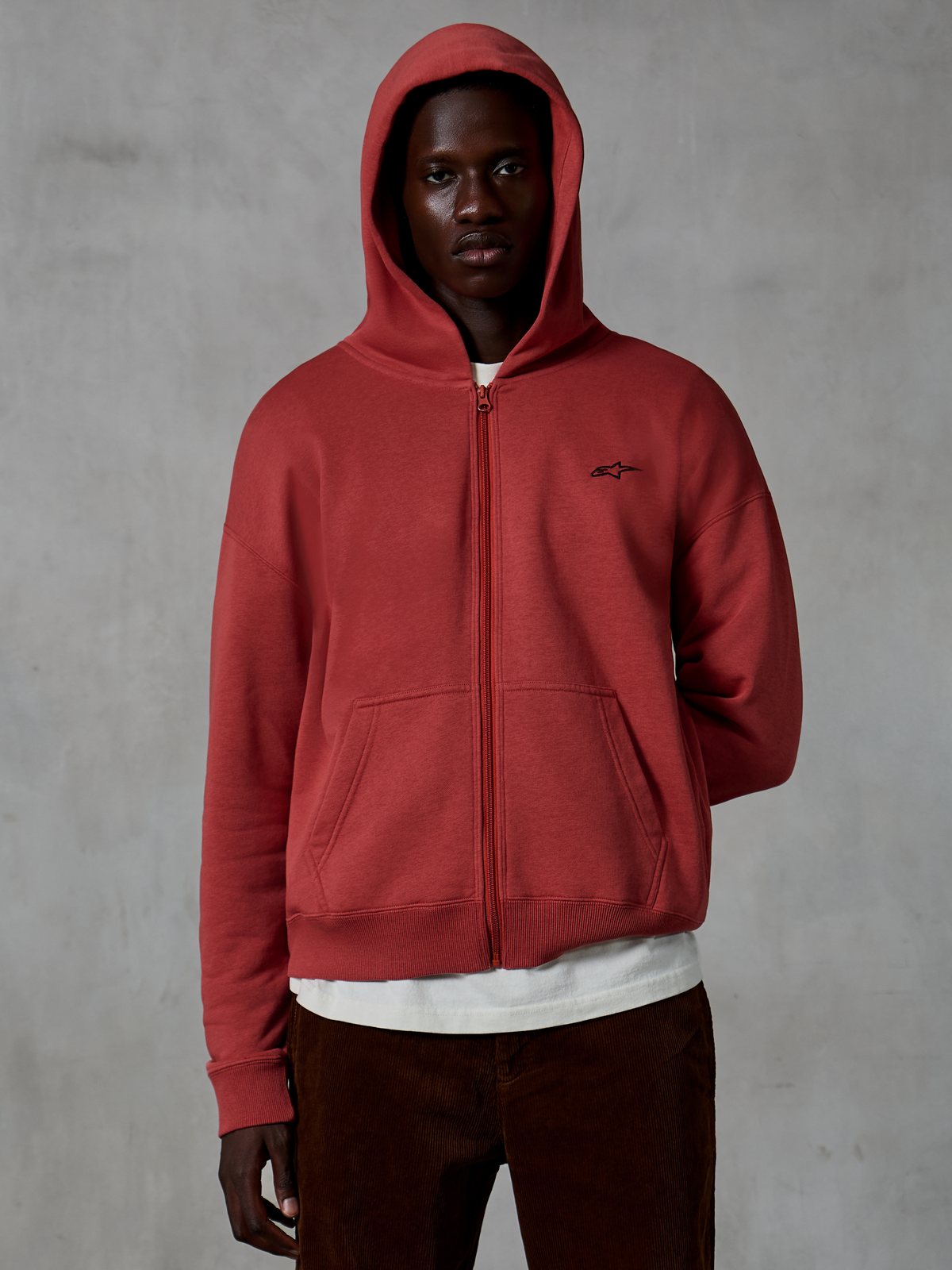 Sudadera con capucha y cremallera Alpinestars Essentials Ageless, ropa deportiva casual en color rojo Brick, diseño de cremallera completa con la capucha puesta, con un pequeño logotipo negro en el pecho, usada por un modelo con camiseta blanca y pantalones de pana marrones sobre un fondo gris.