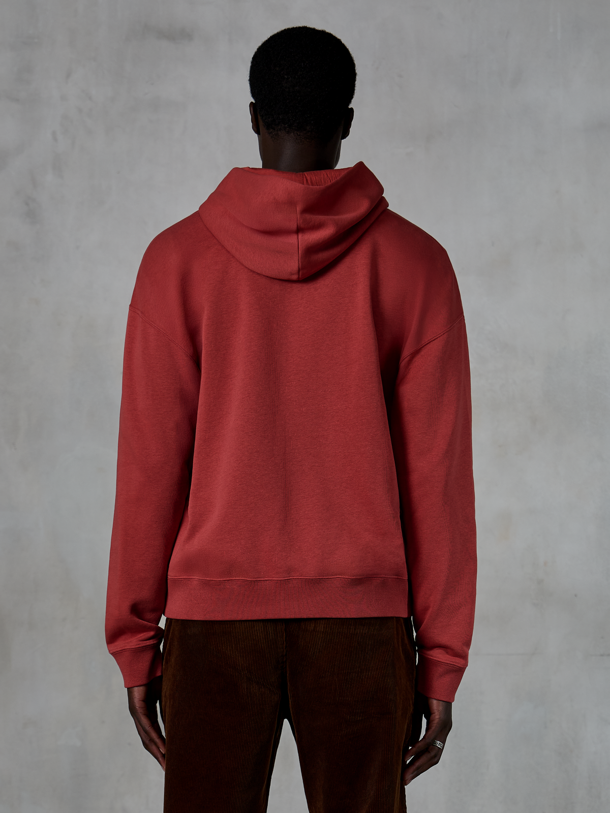 Sudadera con capucha y cremallera Alpinestars Essentials Ageless, rojo ladrillo, vista trasera que muestra el ajuste relajado y la capucha con cordón, combinada con pantalones de pana marrones, ropa urbana casual de inspiración motera