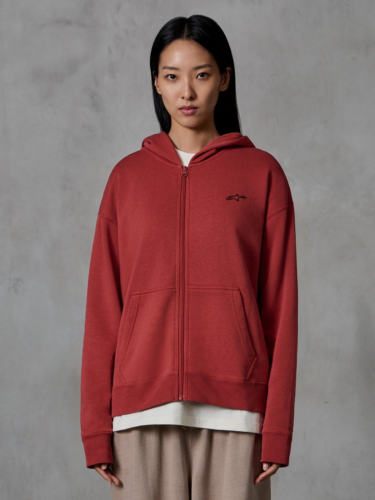 Sudadera con capucha y cremallera Alpinestars Essentials Ageless, color rojo ladrillo, ropa urbana casual, usada por una modelo, diseño con cremallera completa y capucha, pequeño logotipo negro de Alpinestars en el pecho, forro polar compacto de ajuste relajado