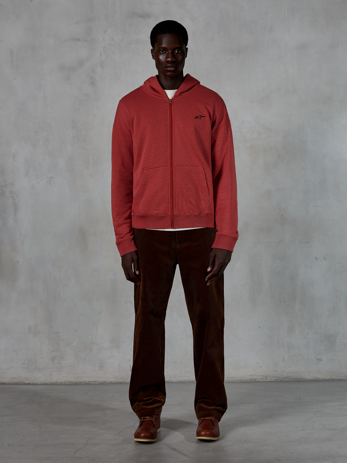 Alpinestars Essentials Ageless Zip Hoodie, ropa urbana casual, rojo ladrillo con pequeño logo de estrella negro, cremallera frontal completa, usada por un modelo con pantalones de pana marrones y botas, diseño premium inspirado en el motociclismo