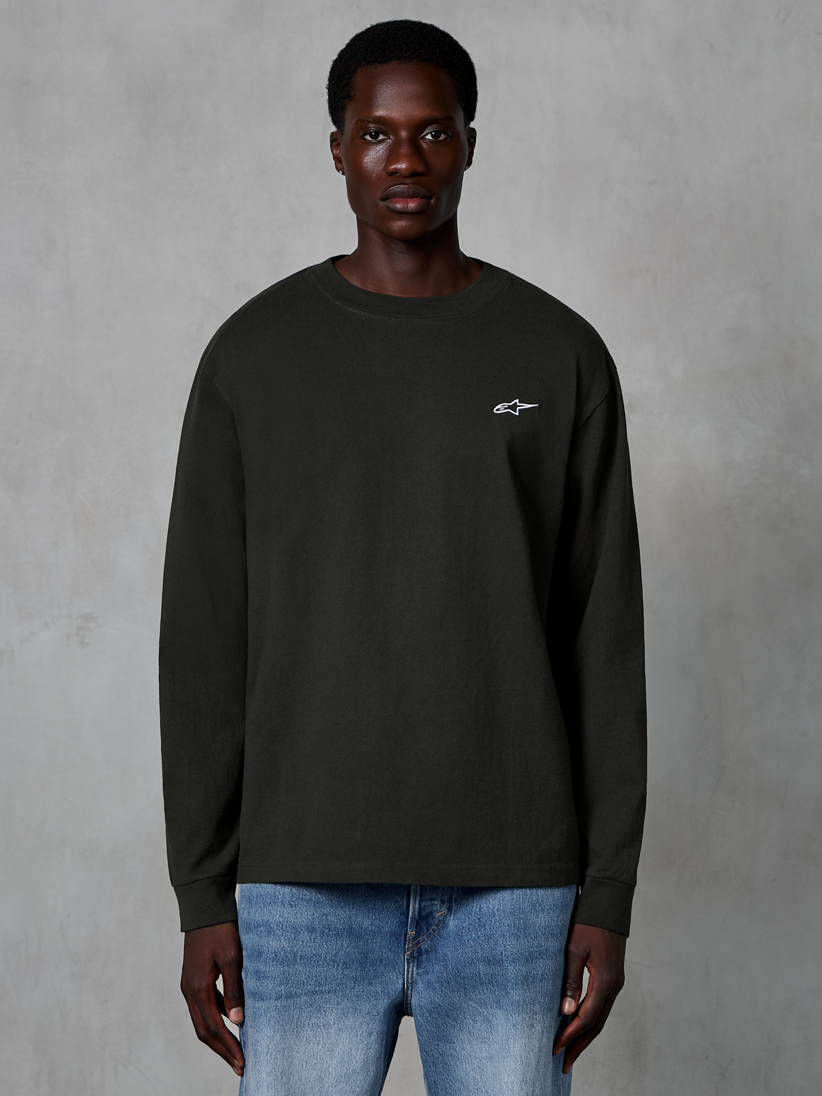 Alpinestars Essentials Ageless LS Tee, camiseta de manga larga, negro Off Black, con un pequeño logo blanco de Alpinestars en el pecho, lucida por un modelo con vaqueros azules, ropa urbana casual de algodón orgánico