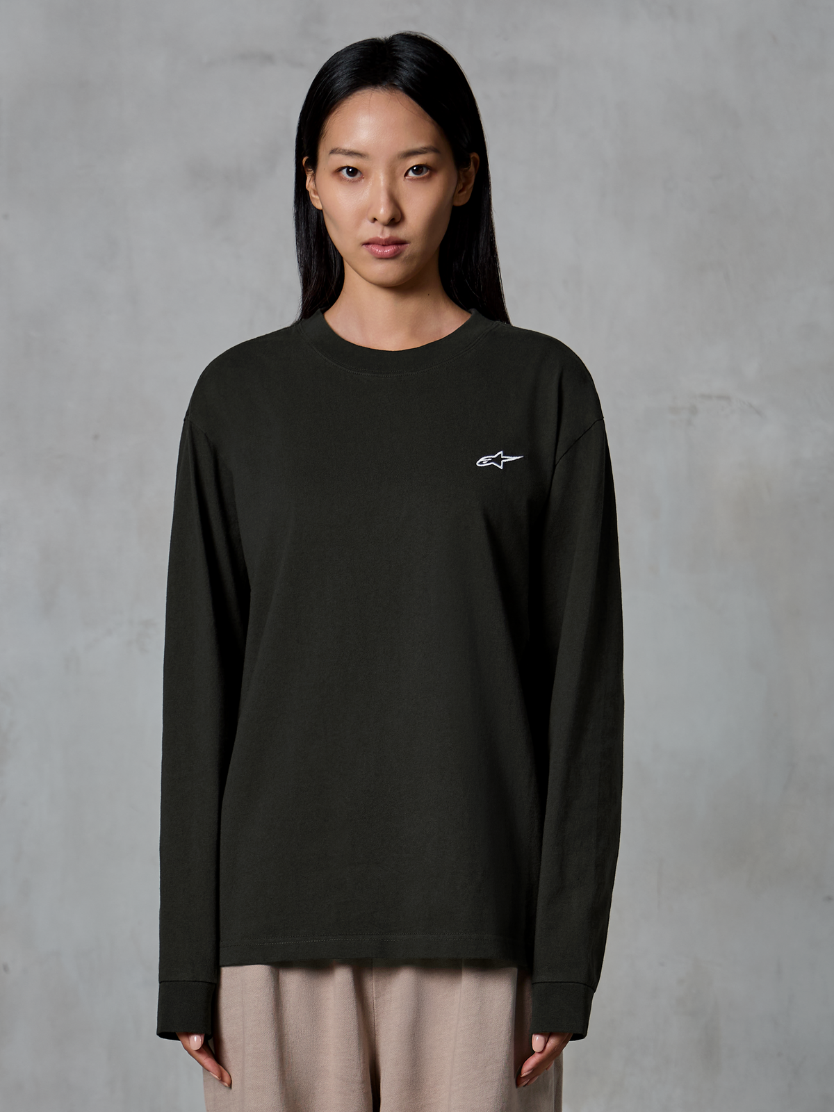 Modelo con la camiseta Alpinestars Essentials Ageless LS Tee, camiseta de manga larga, color negro (Off Black), con un pequeño logo blanco de Alpinestars en el pecho, combinada con pantalones beige, estilo urbano casual