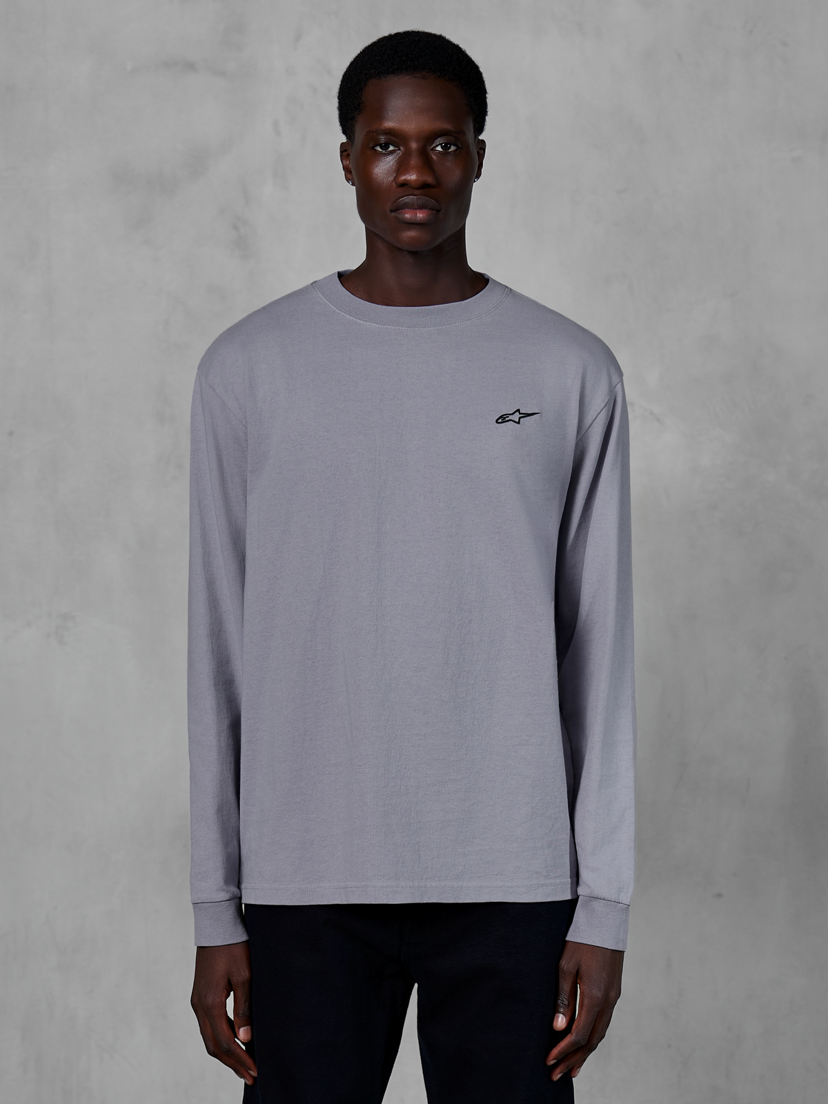 Alpinestars Essentials Ageless LS Tee, camiseta de manga larga, color gris plata, usada por un modelo, con un pequeño logotipo de Alpinestars negro en el pecho, ajuste contemporáneo relajado, estilo urbano casual de algodón orgánico