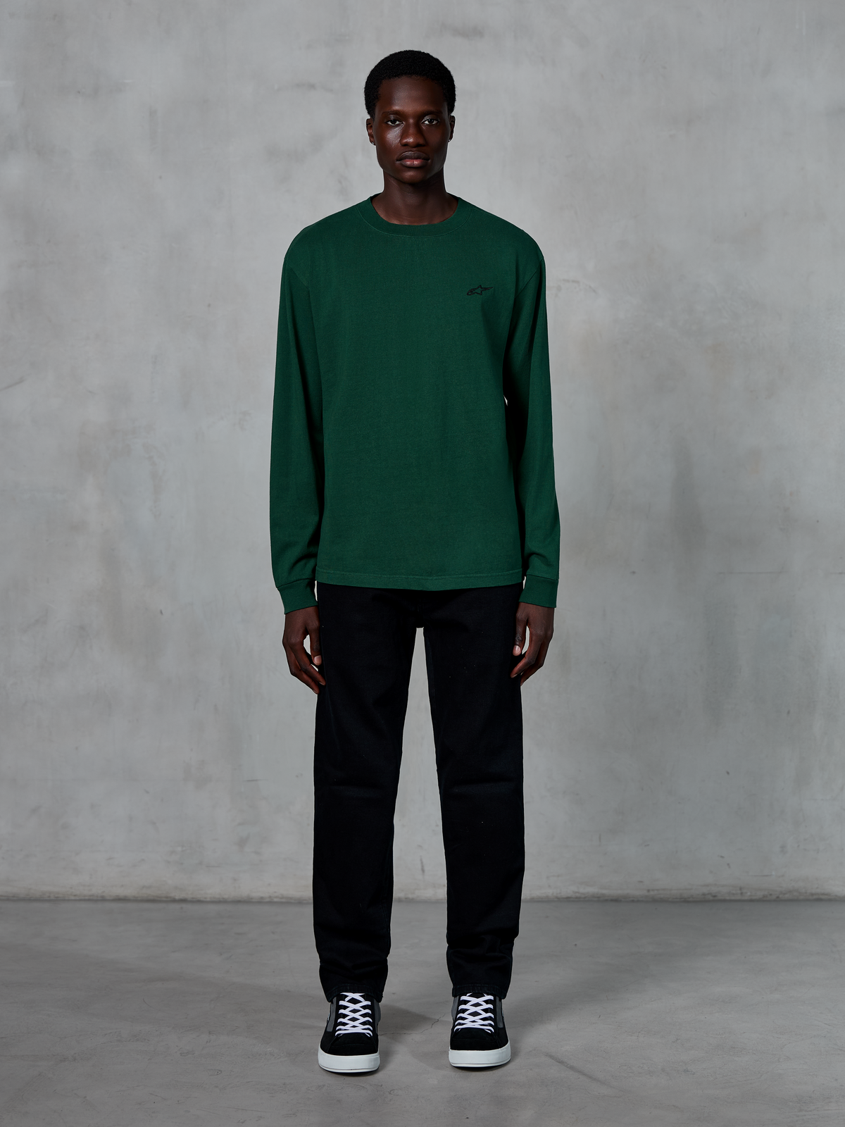 Alpinestars Essentials Ageless LS Tee, camiseta de manga larga, color verde pino, con un pequeño logotipo negro en el pecho, lucida por un modelo con pantalones negros y zapatillas, estilo streetwear casual