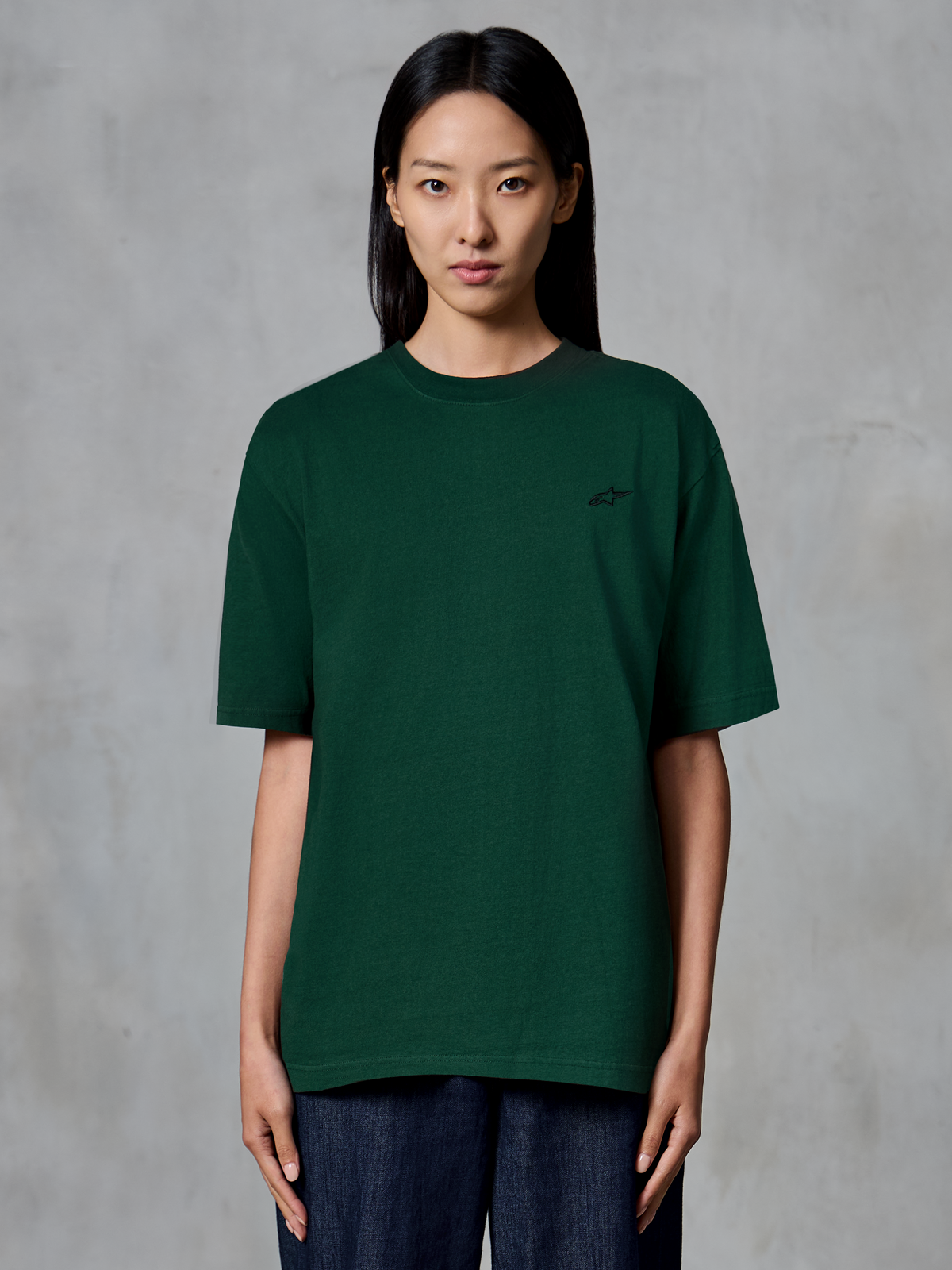 Camiseta Alpinestars Essentials Ageless SS, camiseta de manga corta, verde pino, usada por una modelo, con un pequeño logotipo negro en el pecho, ajuste relajado, moda urbana casual de algodón orgánico