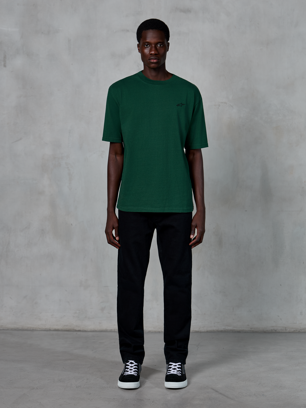 Camiseta Alpinestars Essentials Ageless SS, camiseta de manga corta, verde pino, con un sutil logotipo negro en el pecho, lucida por un modelo con pantalones negros y zapatillas, estilo urbano casual con un ajuste relajado