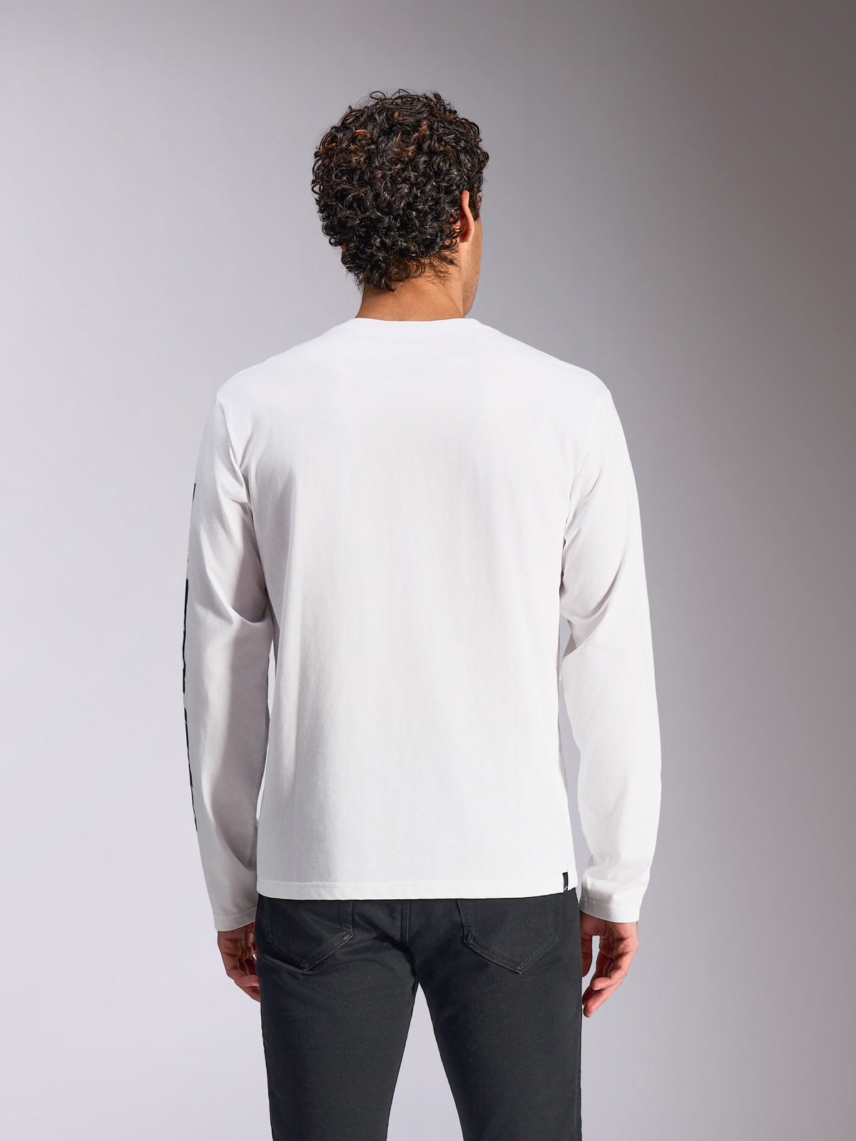 Camiseta Alpinestars Trackside CSF de manga larga, blanca, vista trasera del modelo, combinada con pantalones negros, tejido blanco de manga larga, estilo urbano casual inspirado en el motorsport