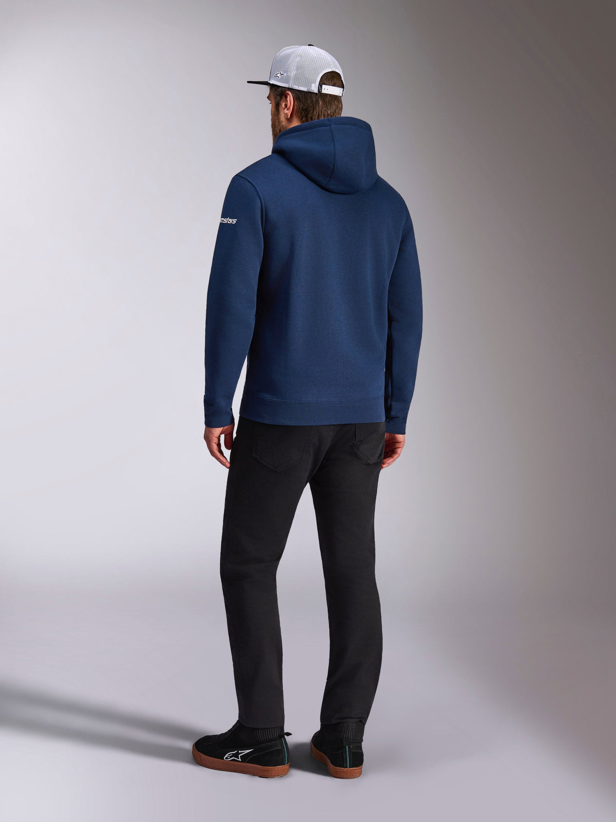 Alpinestars Frontal Hoodie, vista posterior de una sudadera de forro polar en azul marino, logotipo blanco en la manga, combinada con pantalones negros y una gorra trucker de malla blanca, ropa urbana casual inspirada en el automovilismo