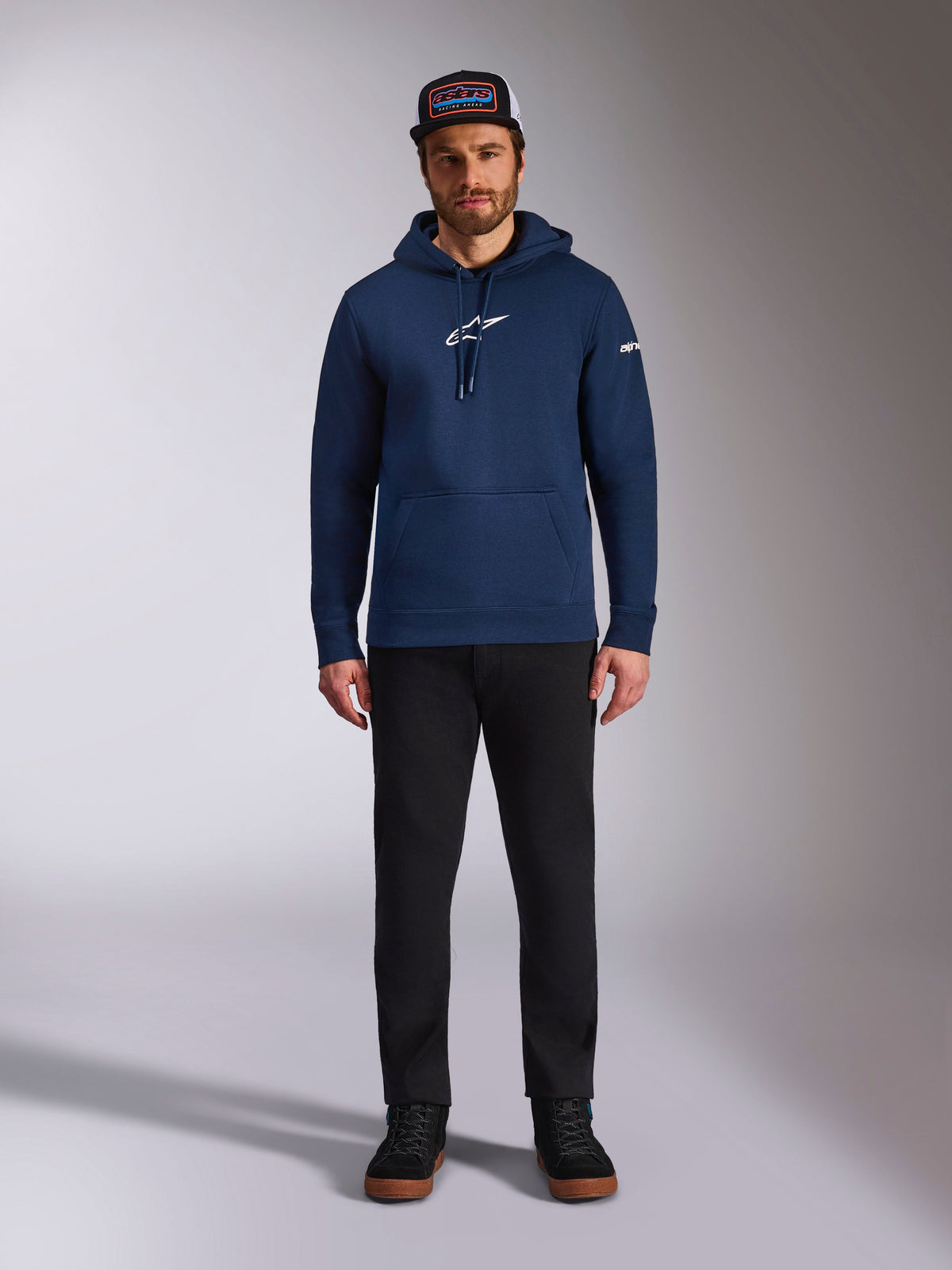 Modelo masculino con la sudadera Alpinestars Frontal Hoodie en color azul marino, sudadera de forro polar con logotipo Astars blanco en el pecho y logo de palabra en la manga, con bolsillo canguro y capucha con cordón, combinada con pantalones negros y gorra de la marca, estilo casual de motorsport