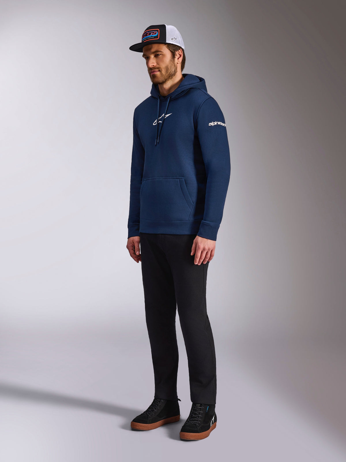 Alpinestars Frontal Hoodie en azul marino, sudadera casual de forro polar con el logotipo de Astars en blanco en el pecho y el logo de palabra en la manga, combinada con una gorra trucker y pantalones negros, ropa de estilo de vida moderna inspirada en el motorsport