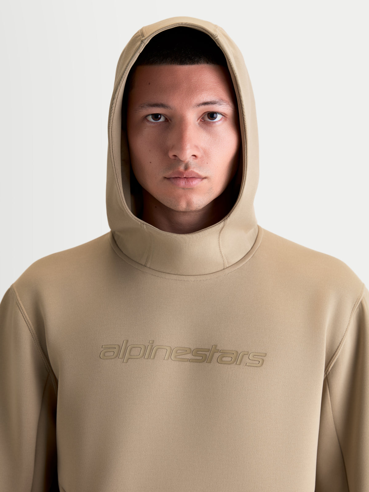 Alpinestars Sudadera con capucha Datrix, pulóver sportswear para hombre y mujer, color marrón, primer plano frontal en modelo con la capucha puesta, logotipo Alpinestars tonal y material de forro polar premium.