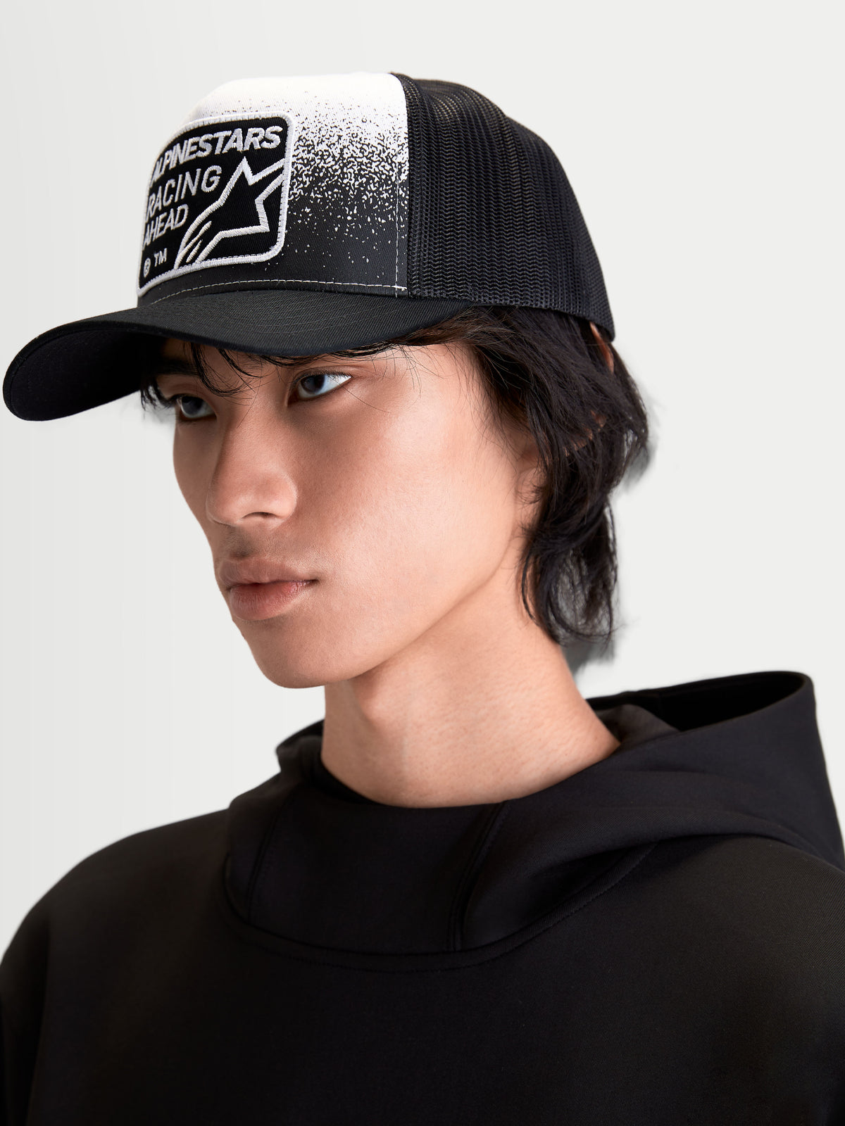 Alpinestars Gravel Trucker Hat, Casual Sportswear Cap on model, Black and White, featuring a structured crown, curved bill, breathable mesh back, and woven patch with gravel-texture graphics and Alpinestars Racing Ahead branding.
