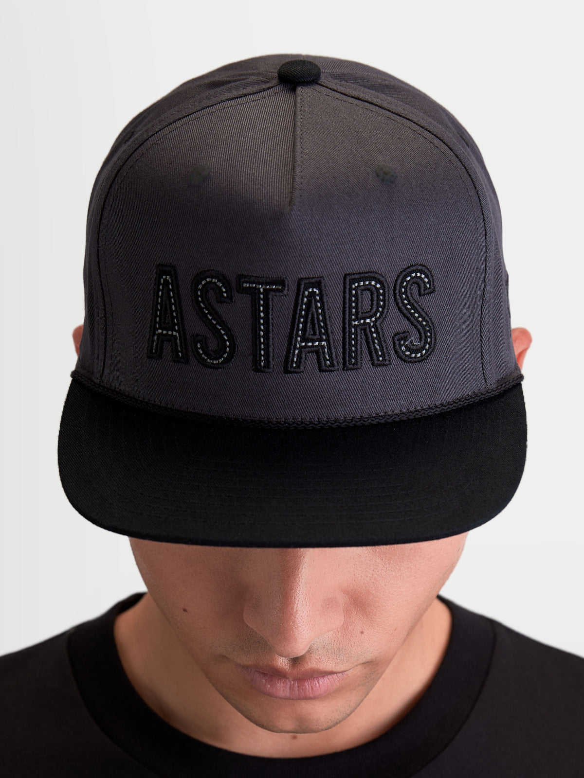 Alpinestars Stadium Snapback, gorra de visera plana estilo motorsport, unisex, gris oscuro y negro, corona estructurada con logotipo ASTARS bordado y costuras en contraste, sarga de algodón, vista frontal en modelo