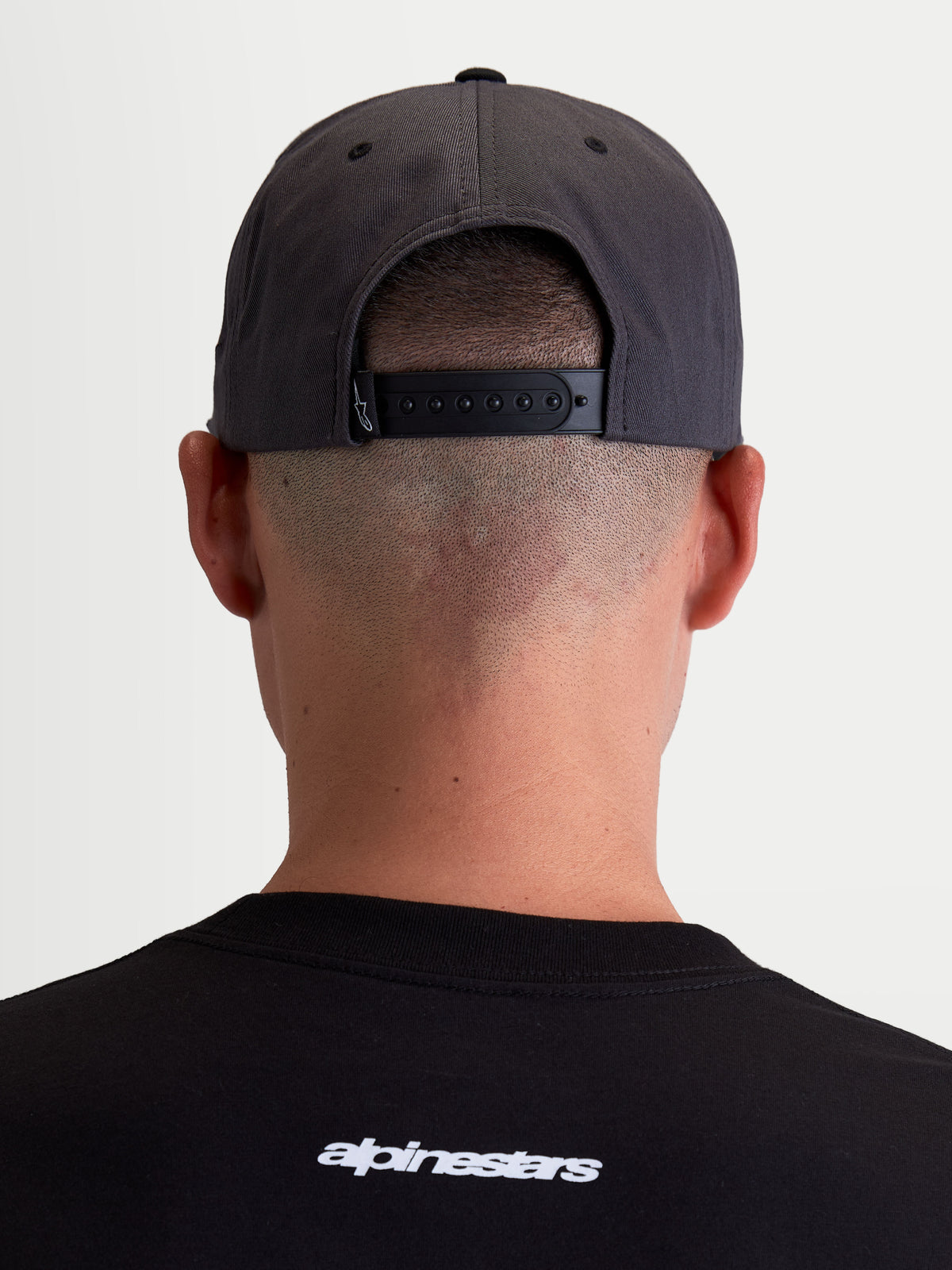 Alpinestars Stadium Snapback, gorra deportiva en gris oscuro y negro, vista trasera en modelo mostrando el cierre snapback ajustable y la corona estructurada, construcción de sarga de algodón resistente.