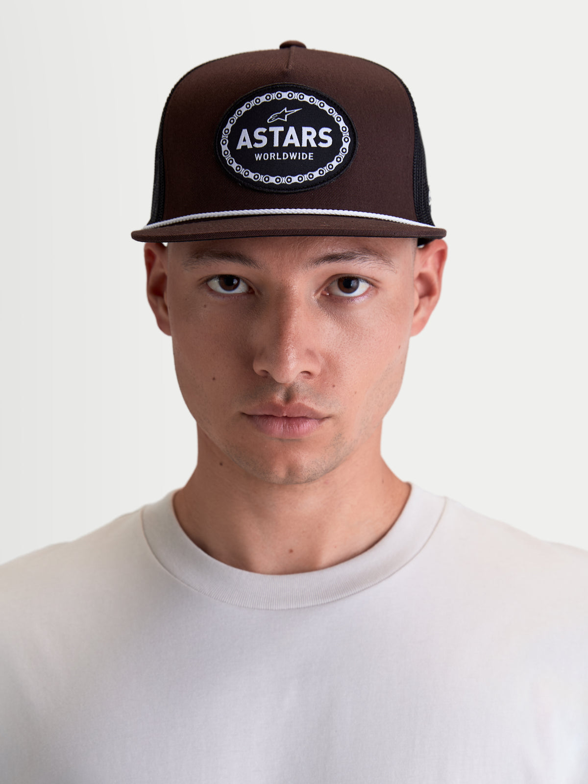 Alpinestars Gorra Hotlink, gorra de estilo sportswear de inspiración motorsport, marrón y negra, con corona estructurada, visera plana con detalle de cordón blanco y parche frontal tejido con el logotipo Astars Worldwide, mostrada en modelo