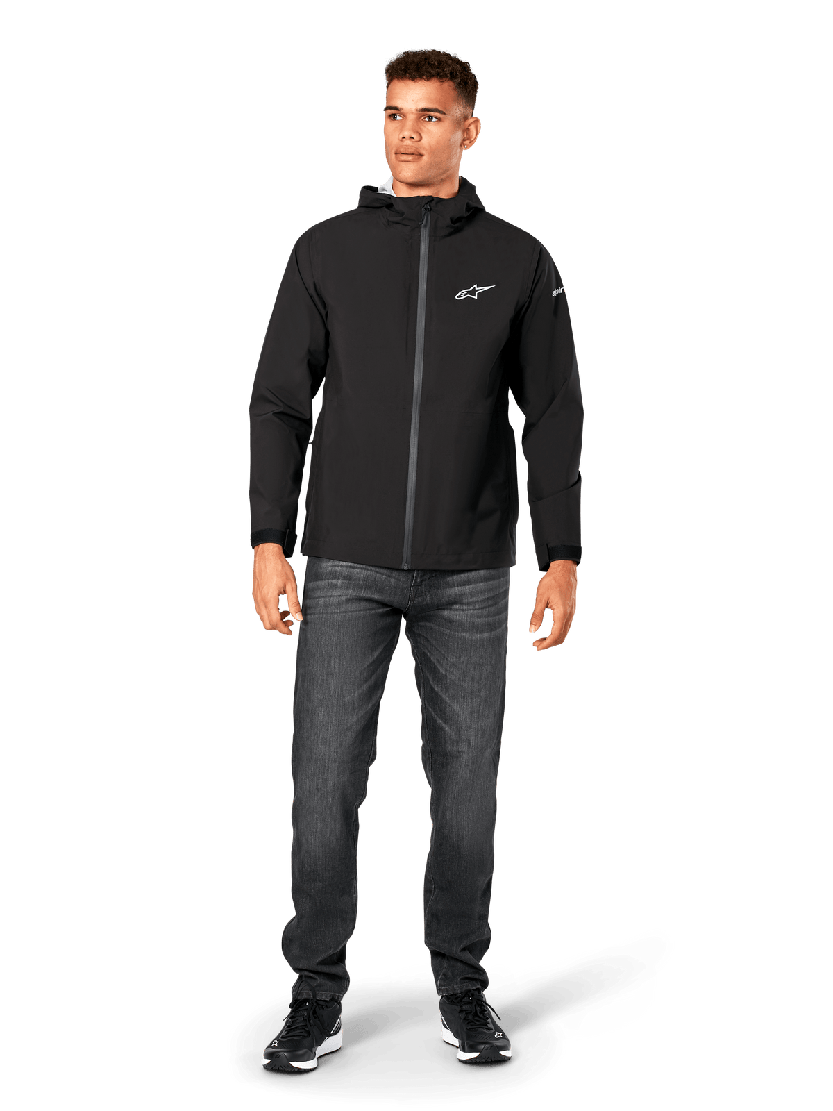 Chaqueta impermeable Alpinestars Kascade, chaqueta de lluvia para hombre, negra, combinada con vaqueros gris oscuro y zapatillas negras, con diseño con capucha, cremallera frontal recubierta y logotipo blanco de Alpinestars en el pecho, ropa urbana casual de inspiración motorsport