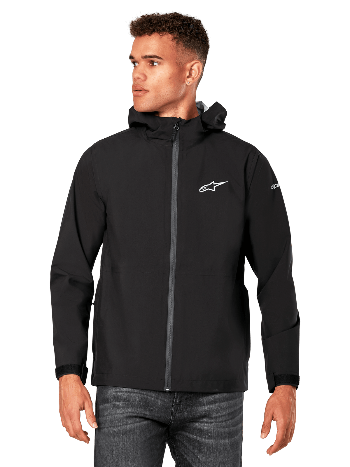 Modelo masculino vistiendo la chaqueta impermeable Alpinestars Kascade, negra, con logotipo blanco en el pecho y marca en la manga, chaqueta de lluvia con capucha, cremallera frontal recubierta y puños de velcro, combinada con vaqueros oscuros, ropa urbana casual inspirada en el automovilismo