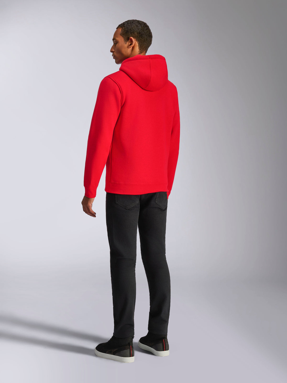 Sudadera con capucha Alpinestars Ageless V3, estilo urbano casual, rojo vibrante, vista trasera del modelo, con capucha con forro de jersey, puños y dobladillo de canalé, combinada con pantalones negros, diseño moderno inspirado en el automovilismo