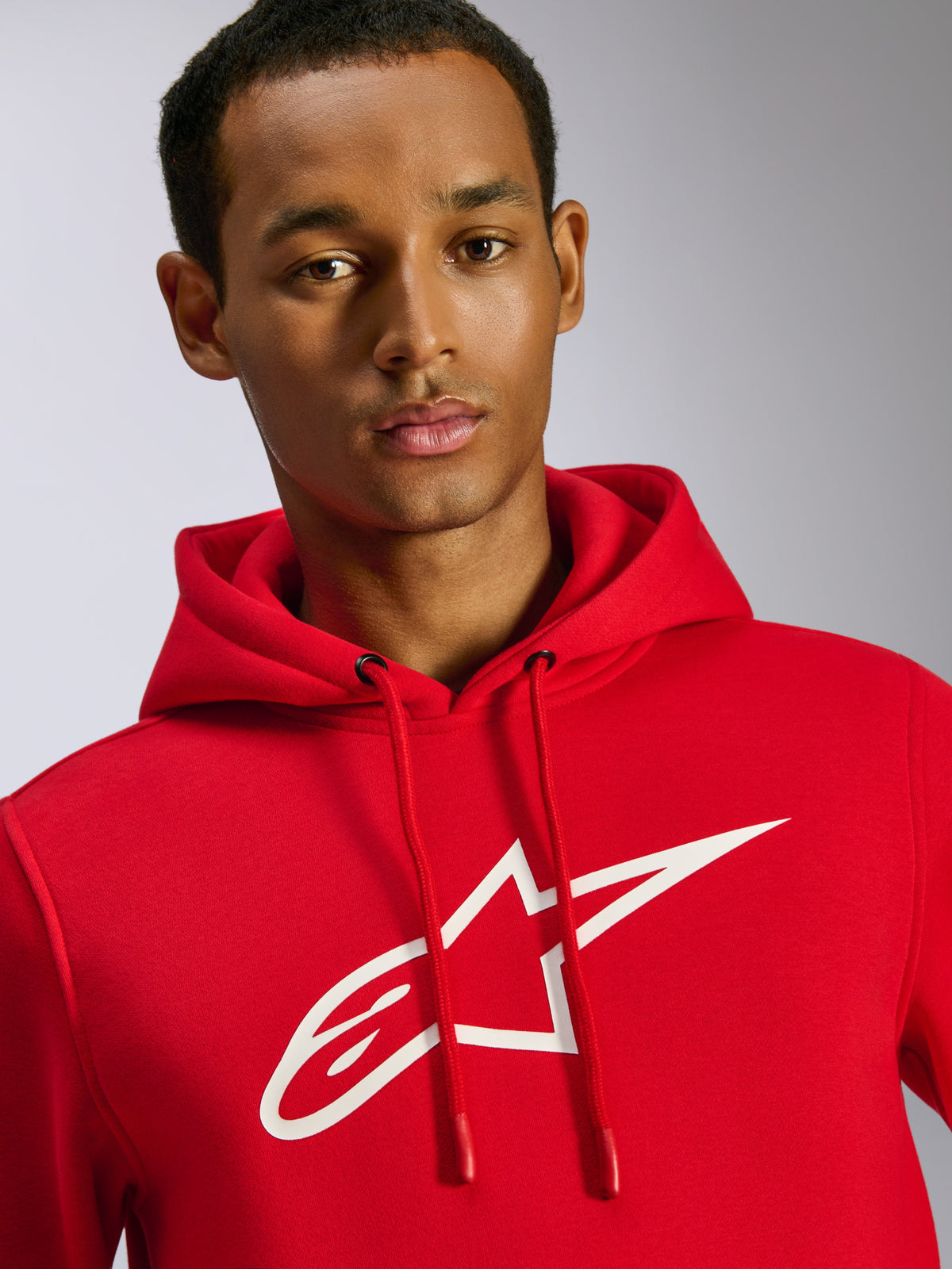 Sudadera con capucha Alpinestars Ageless V3, roja, con un gran logotipo blanco de Alpinestars en el pecho, capucha con cordón y puntas sumergidas, ropa urbana casual inspirada en el automovilismo, vestida por un modelo masculino