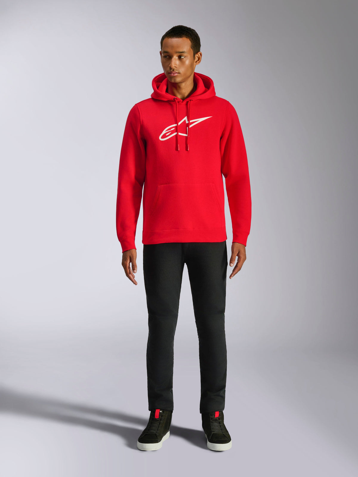 Sudadera con capucha Alpinestars Ageless V3, ropa urbana casual, roja con el logotipo de Alpinestars bordado en blanco, lucida por un modelo masculino con pantalones negros, capucha con cordón, bolsillo de canguro delantero, diseño moderno inspirado en el mundo del motor