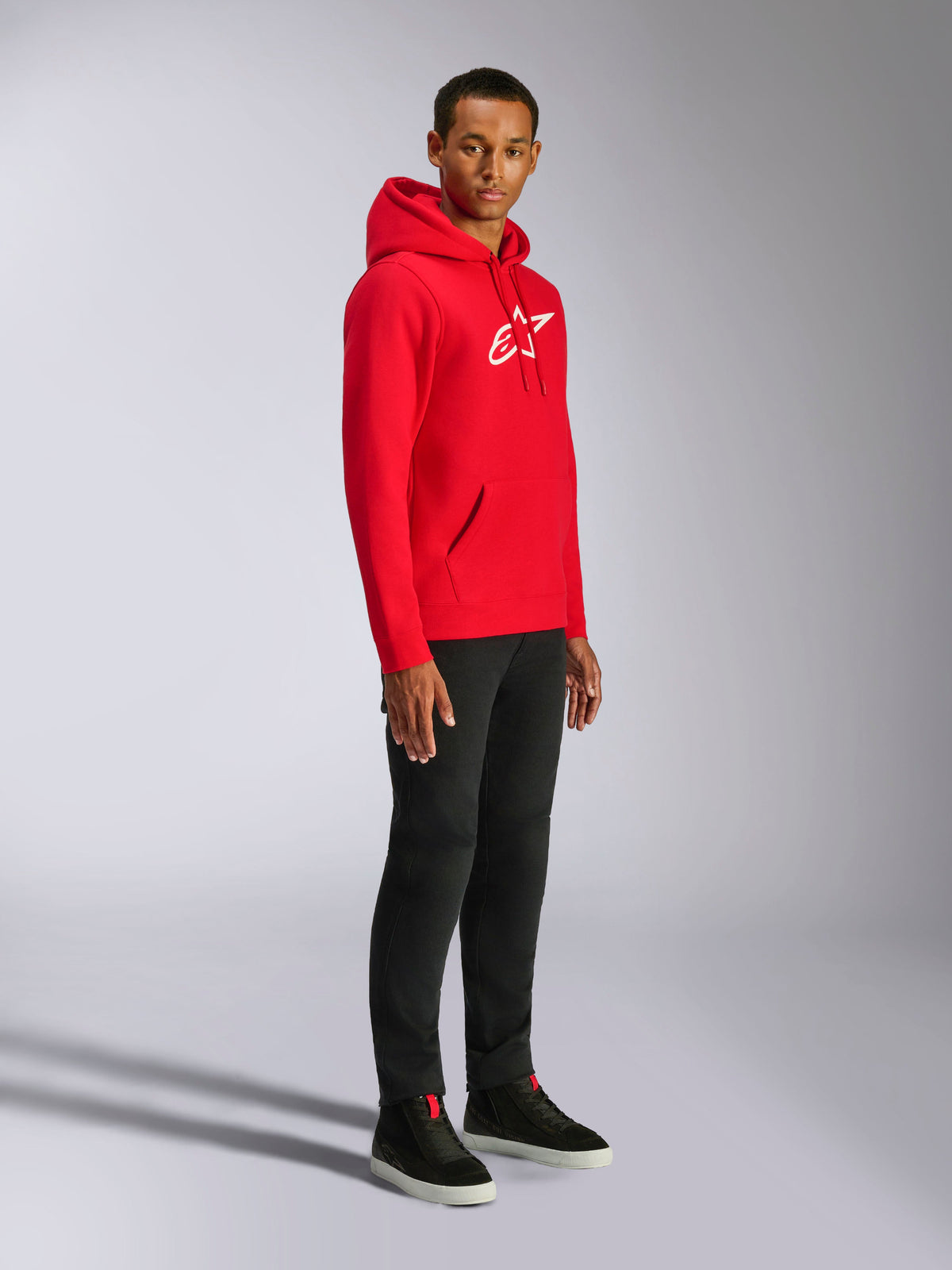 Alpinestars Ageless V3 Hoodie, roja con logotipo bordado en blanco, sudadera deportiva casual lucida por un hombre con pantalones negros y zapatillas negras, con capucha ajustable mediante cordón y puños de canalé
